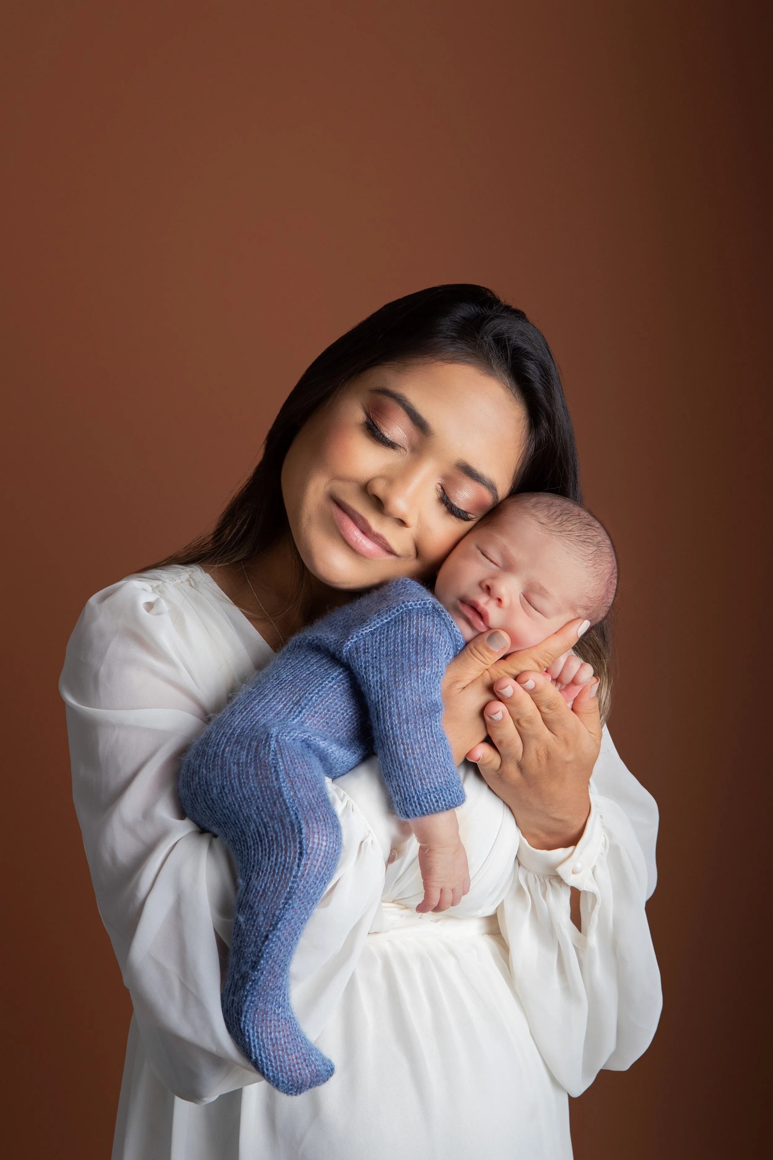 A woman with closed eyes gently holds a sleeping baby dressed in a blue sweater, both embracing affectionately against a brown background.