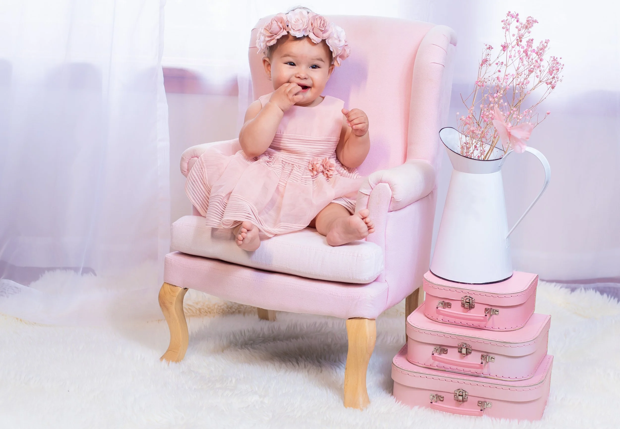 A young girl sitting on a pink armchair wearing a pink dress and a floral headband, with pink suitcases and a white pitcher with pink flowers beside her.