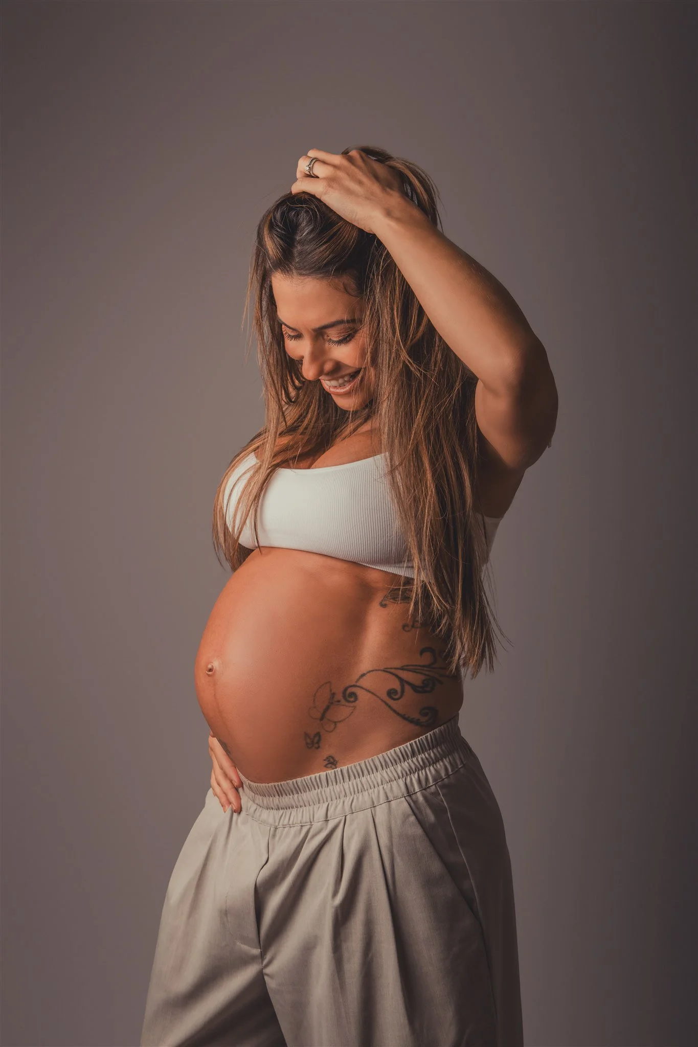 A pregnant woman smiling with her eyes closed, wearing a white sports bra and beige pants, touching her head with one hand and her belly with the other, against a plain gray background.