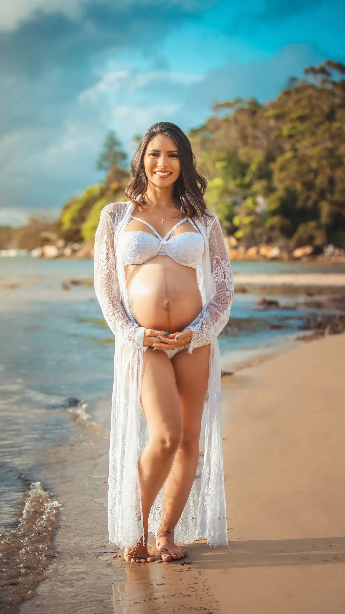 A pregnant woman in white lingerie and a lace cover-up standing on a beach with trees and blue sky in the background.