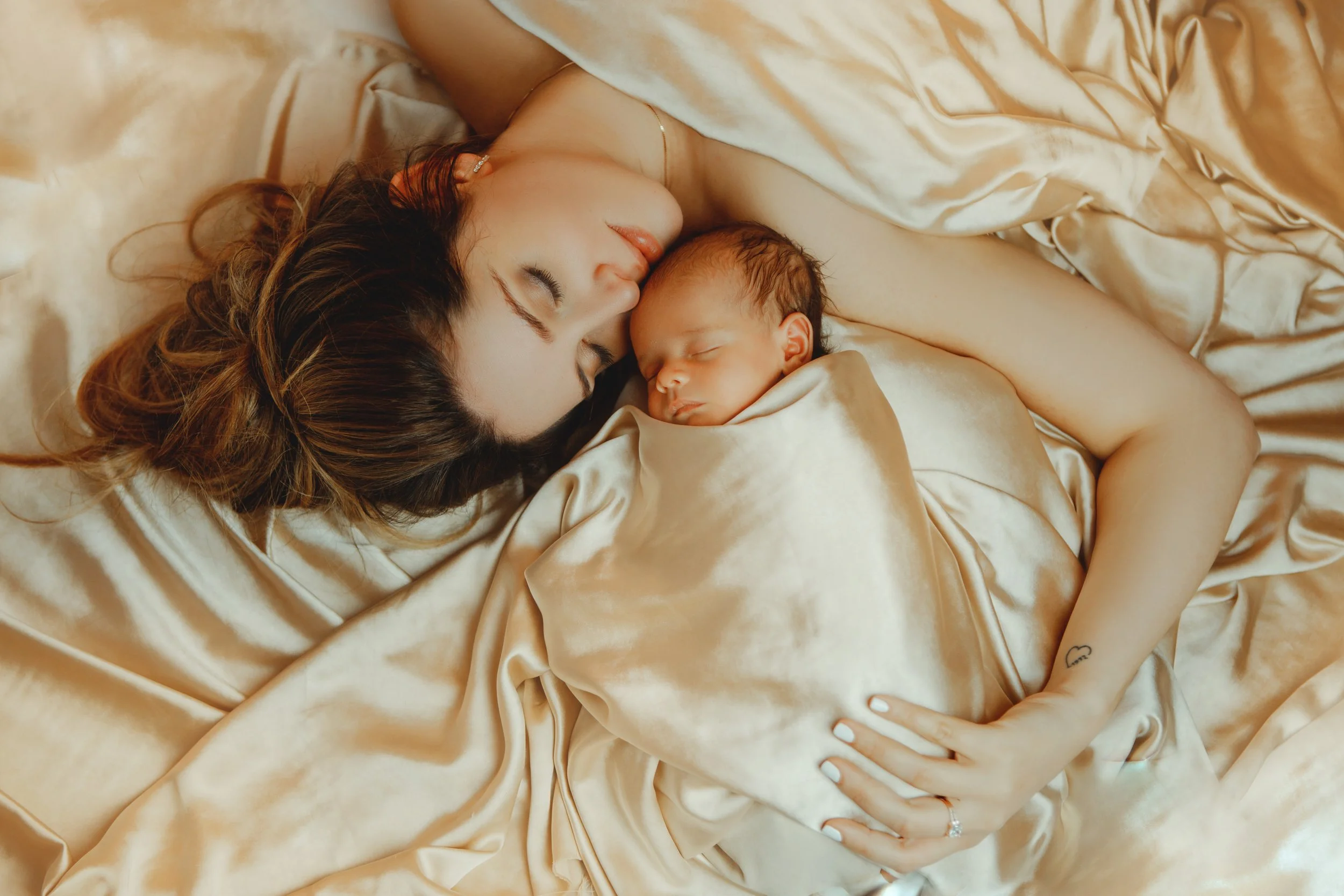 A woman and a newborn baby sleeping together on a satin sheet, the woman with long brown hair and a small heart tattoo on her wrist.