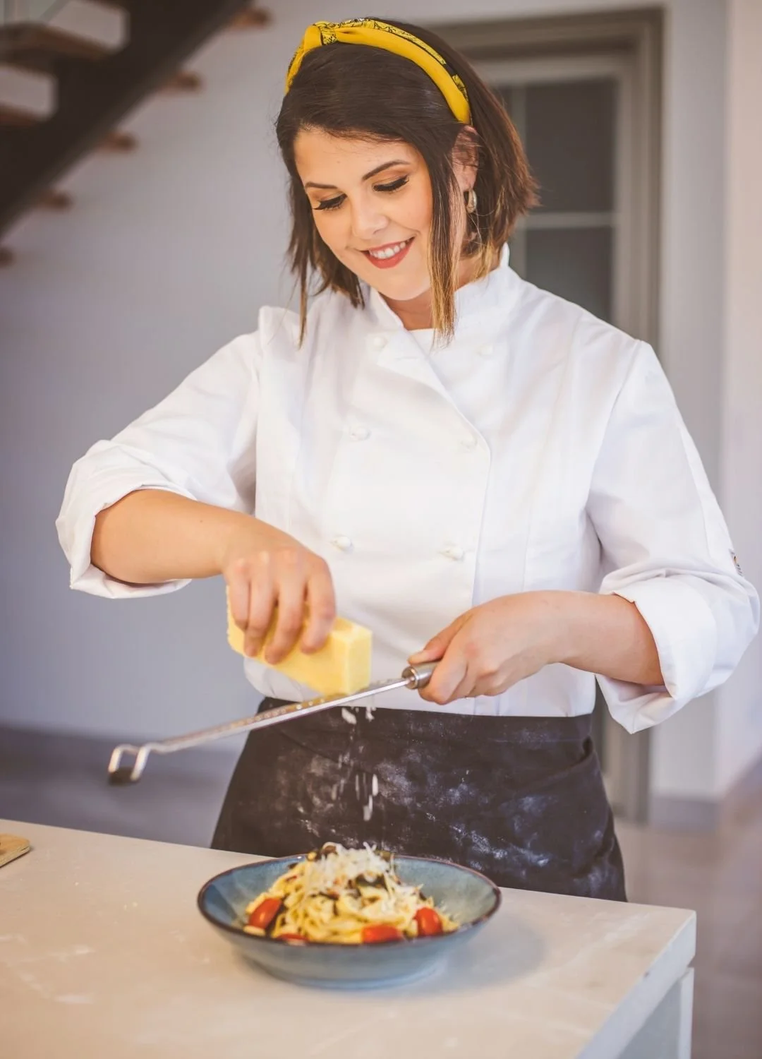 A woman in a white chef's coat and yellow headband grating cheese over a plate of pasta with tomatoes.