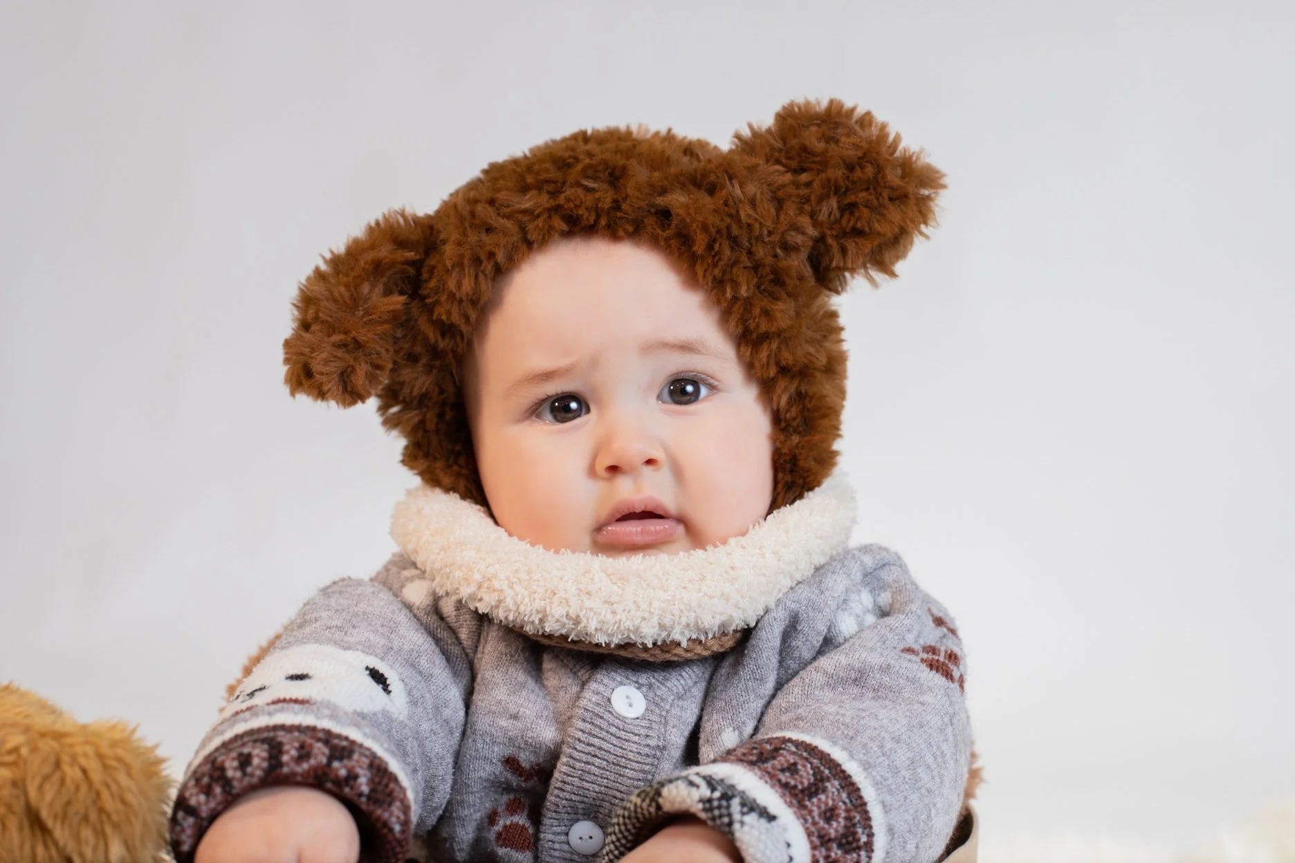Baby wearing a brown teddy bear hat and cozy gray sweater, looking surprised against a plain light background.