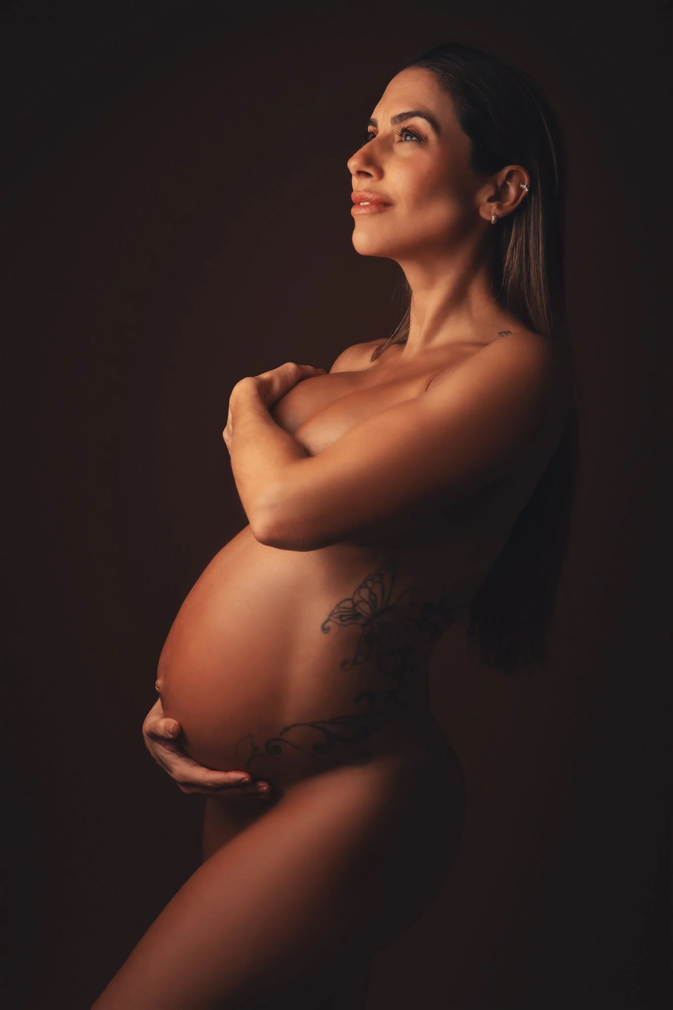 A pregnant woman with long brown hair and tattoos, cradling her belly and looking upward with a serene expression against a dark background.