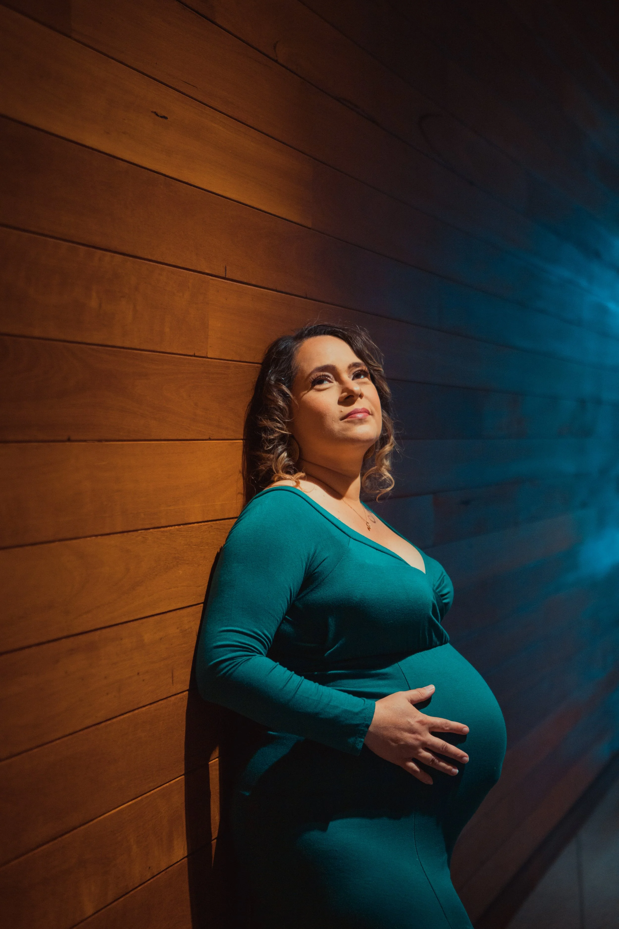 Pregnant woman in a teal dress leaning against a wooden wall, looking confidently at the camera.
