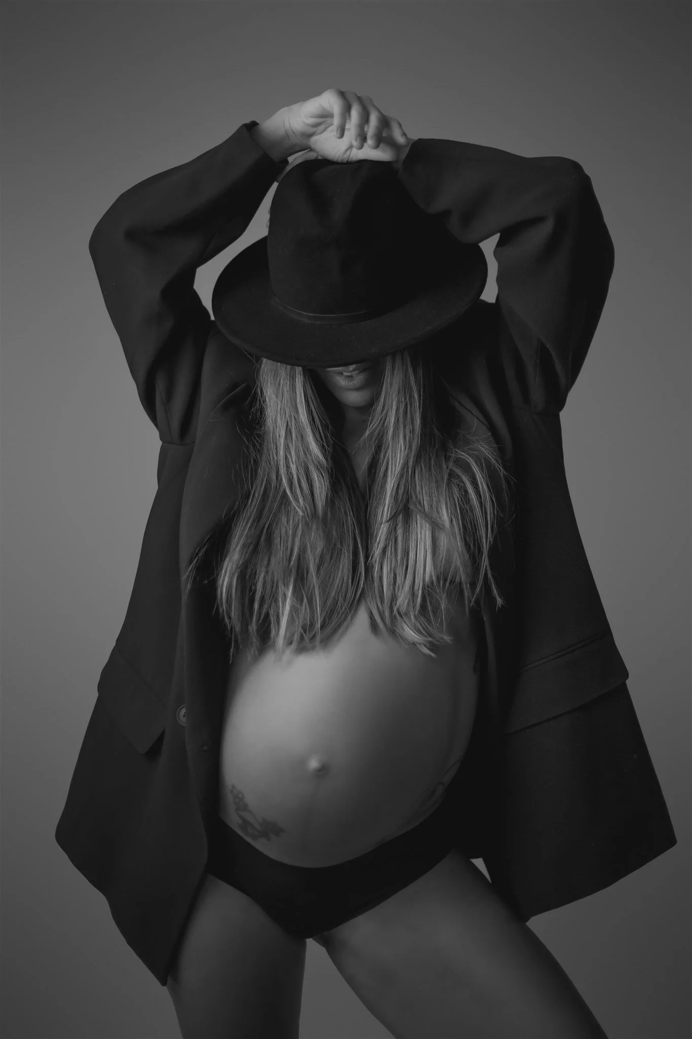 Black and white portrait of a pregnant woman wearing a hat, with her long hair down, a jacket, and underwear, posing with her hands resting on her head.