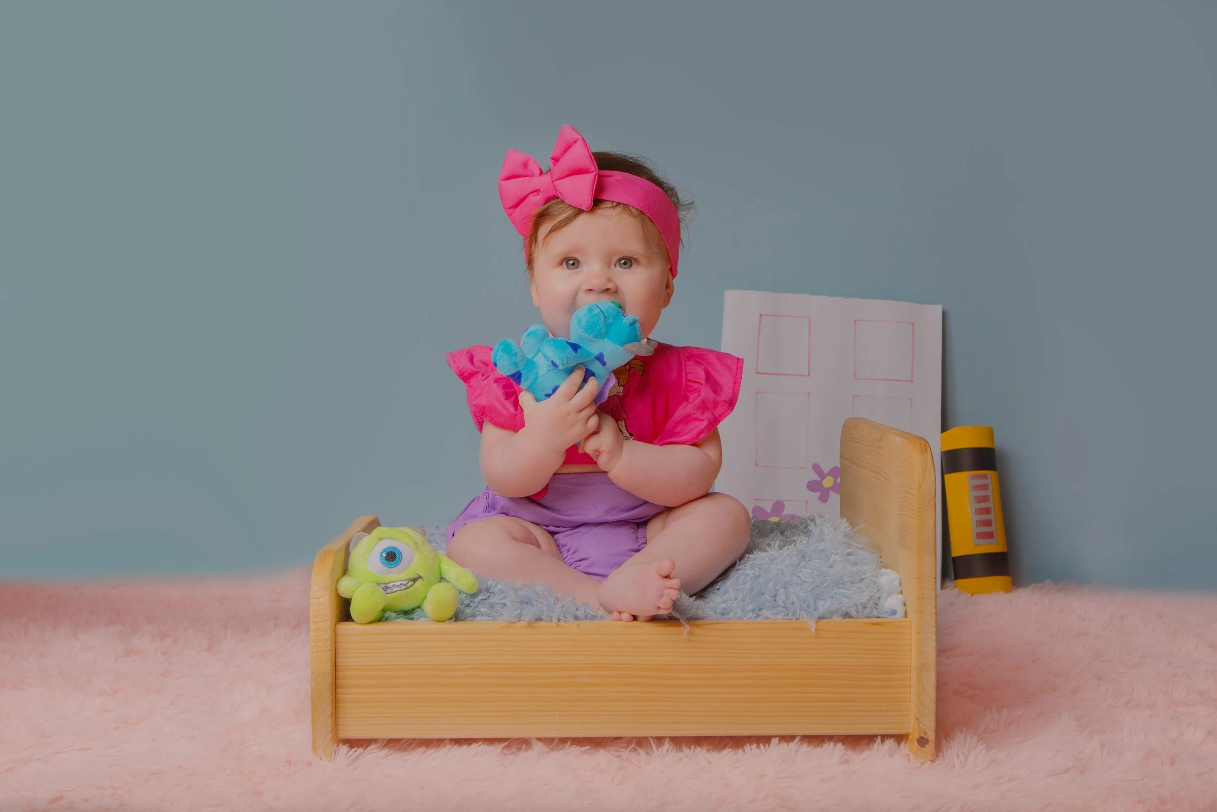 Adorable baby girl with pink bow headband sitting on a small bed, holding a colorful plush toy, surrounded by other toys and drawings, on a soft pink rug.