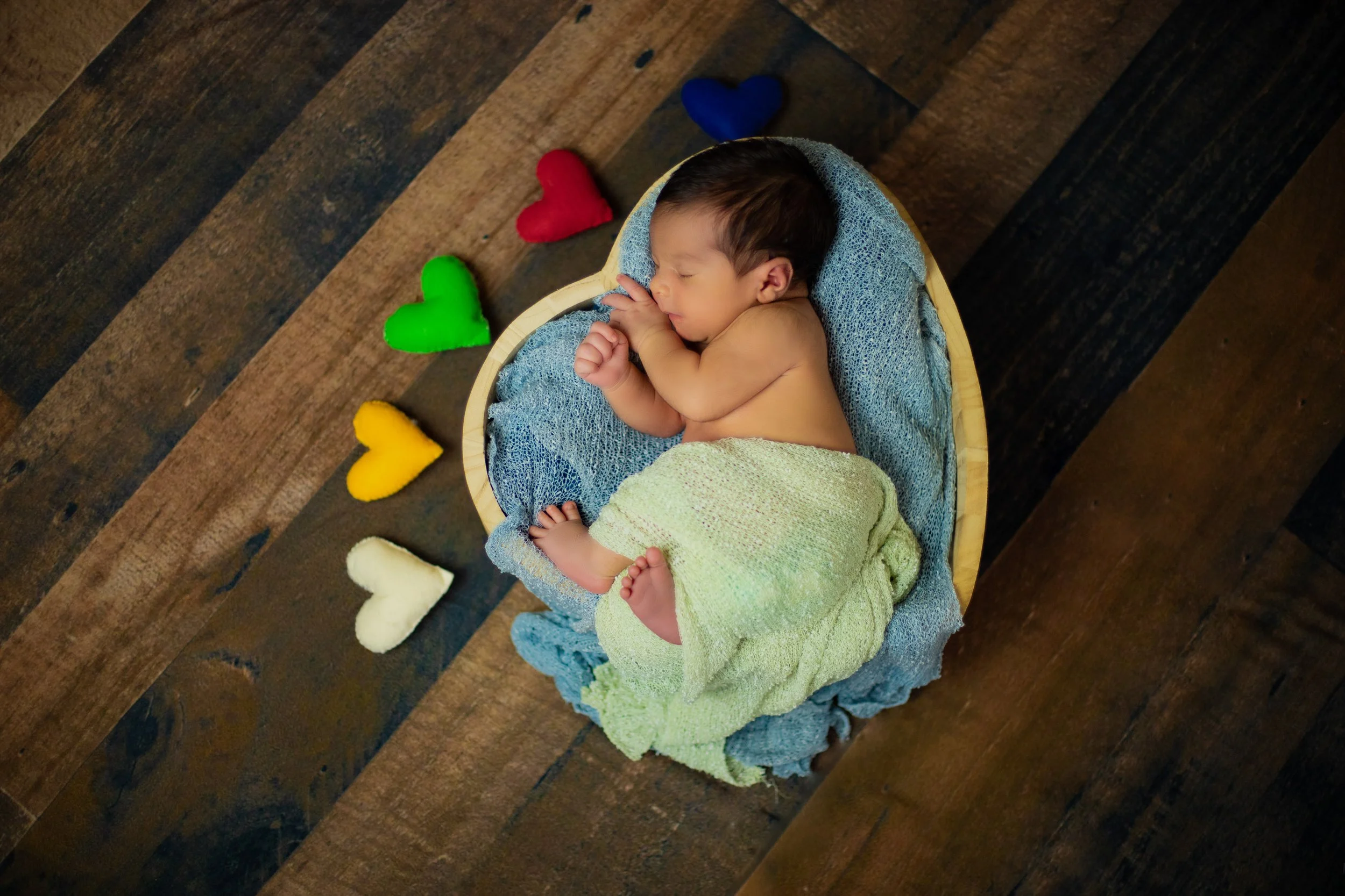 A sleeping baby wrapped in a light green towel, lying on a blue blanket inside a wooden bowl, surrounded by multicolored felt hearts on a wooden floor.