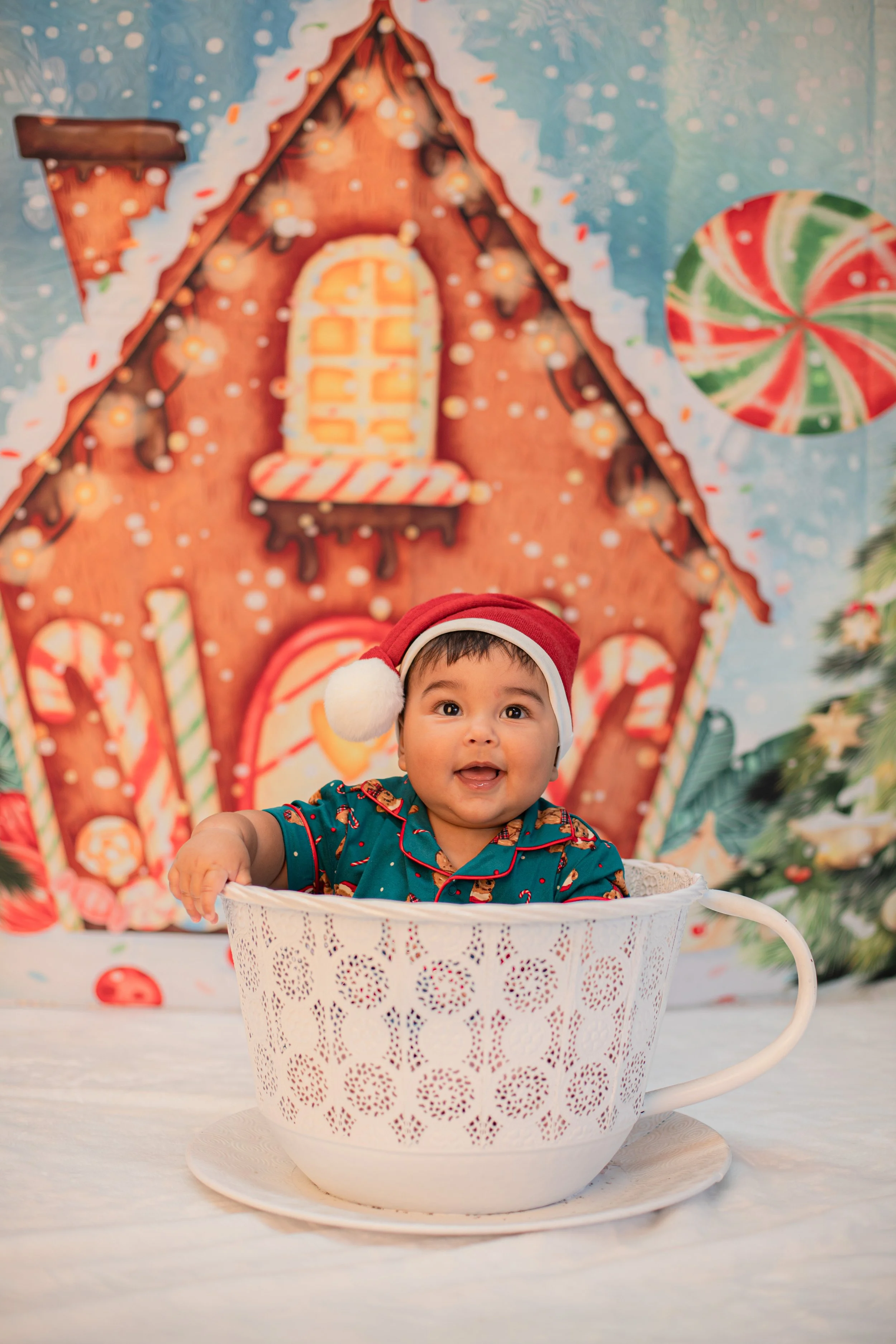 A smiling baby wearing a red Santa hat with white trim, sitting inside a large white decorative cup against a Christmas-themed backdrop featuring a gingerbread house and holiday decorations.