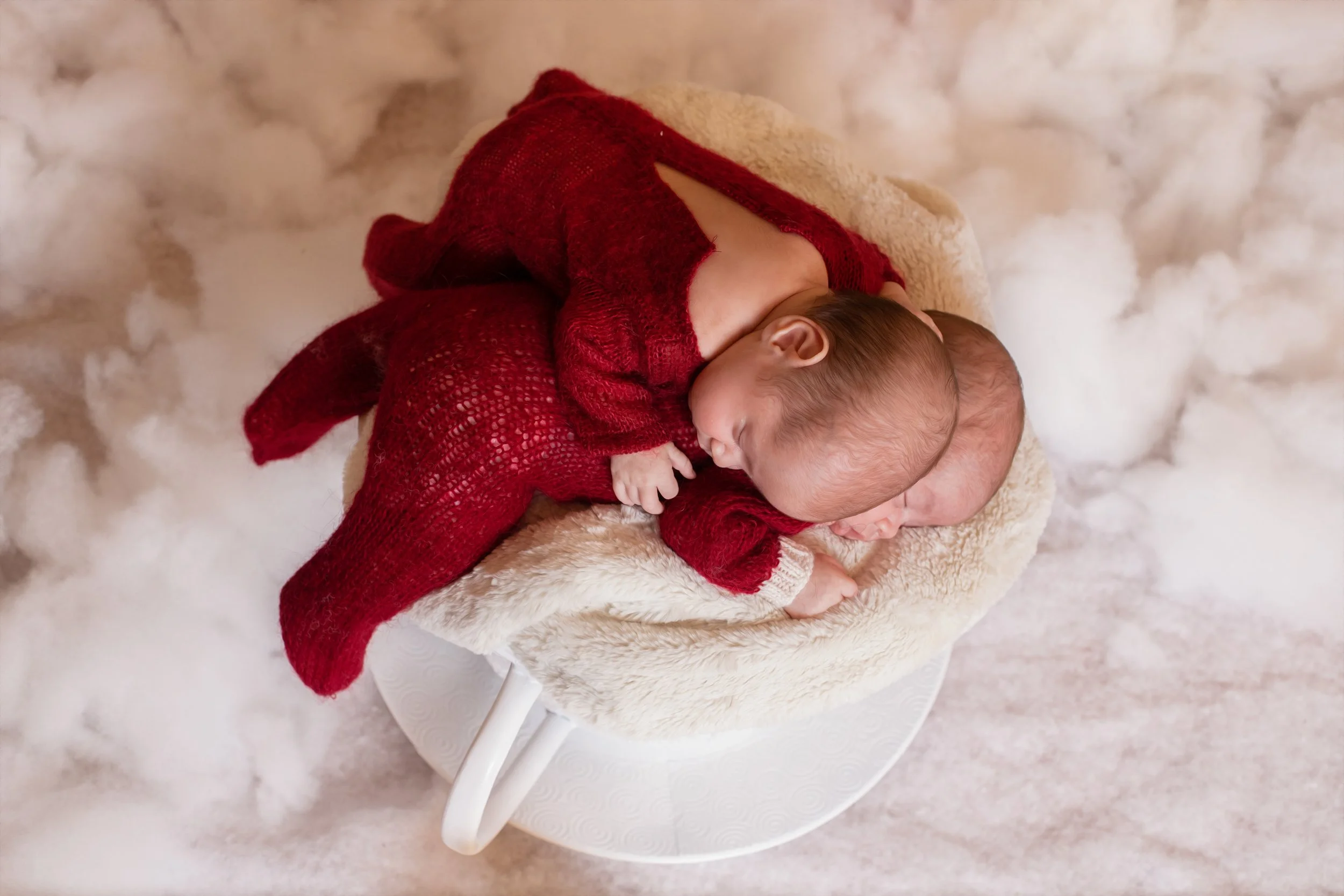 Two newborn babies sleeping together on a soft, furry blanket in a cozy basket, wearing red knitted outfits.