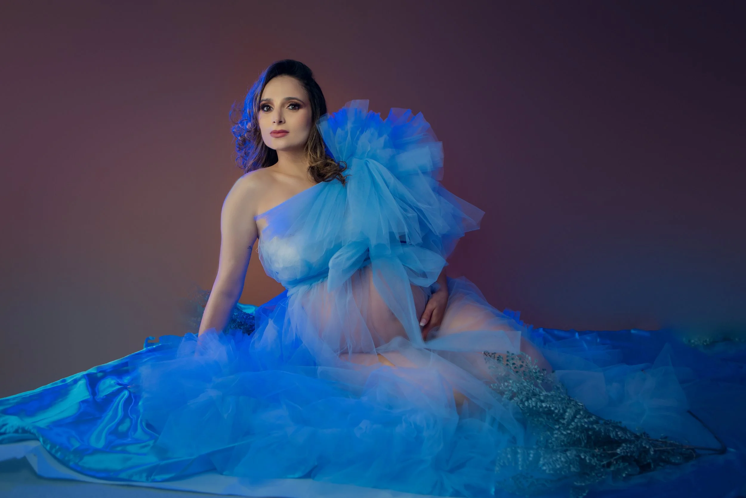 A woman sitting on the floor wearing a flowing blue tulle dress with large ruffles, looking at the camera with a neutral expression, against a gradient background.