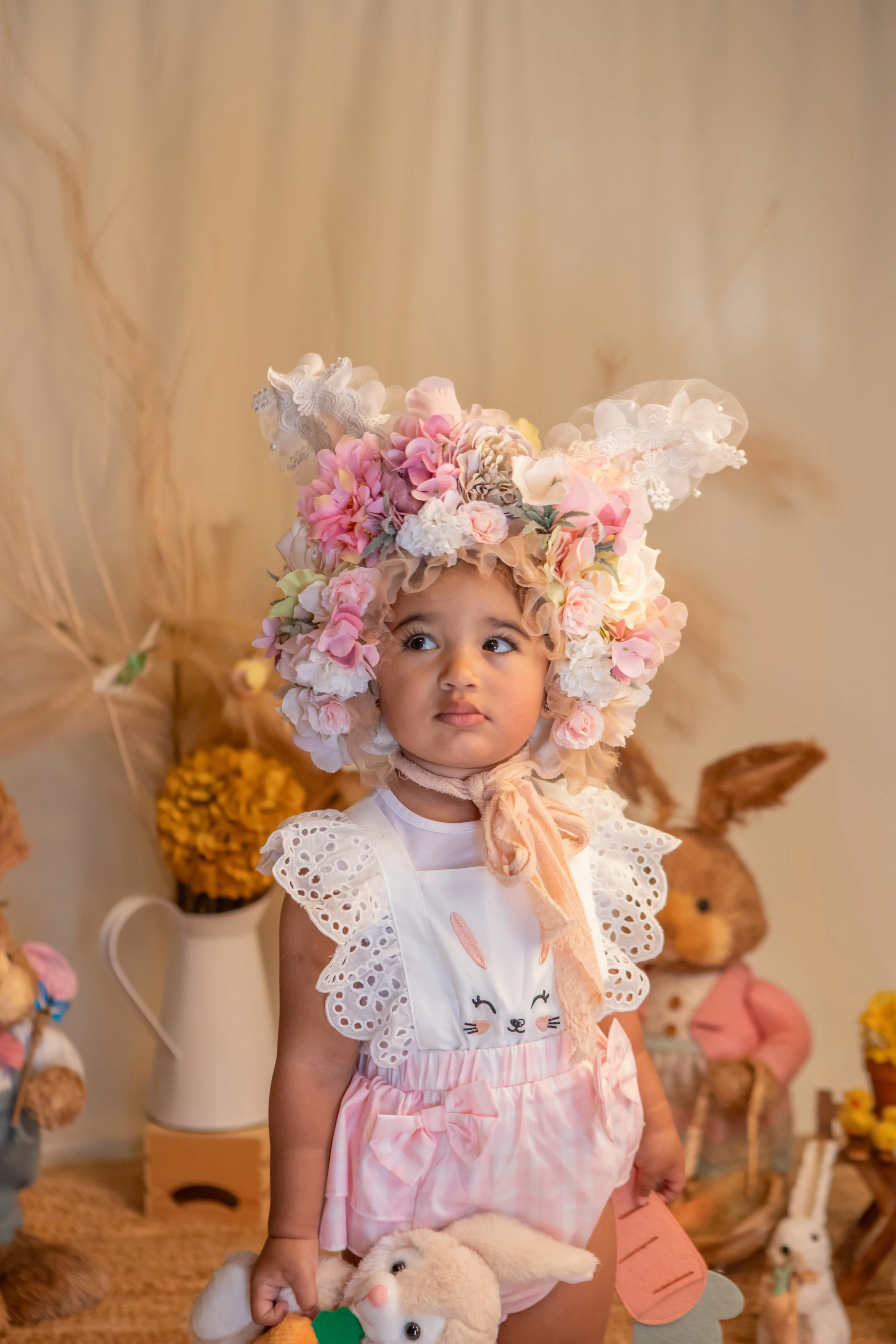 Young girl dressed as a bunny, wearing a floral headpiece and pink and white clothing, surrounded by bunny and animal plush toys and decorations.