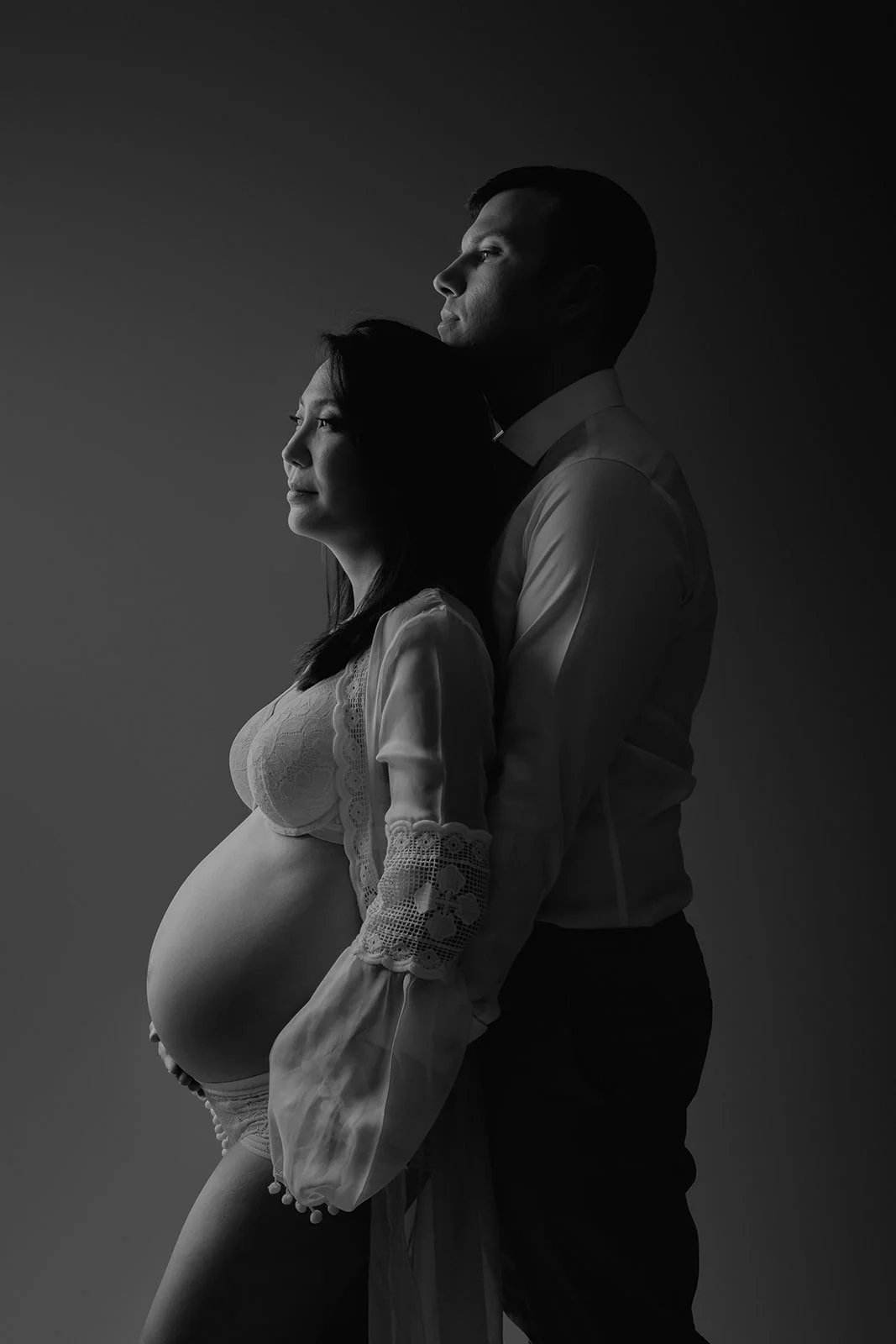 A black and white silhouette of a pregnant woman and a man standing behind her, both facing sideways. The woman is wearing lace lingerie and a sheer robe, highlighting her baby bump. The man is dressed in a button-down shirt. The portrait emphasizes