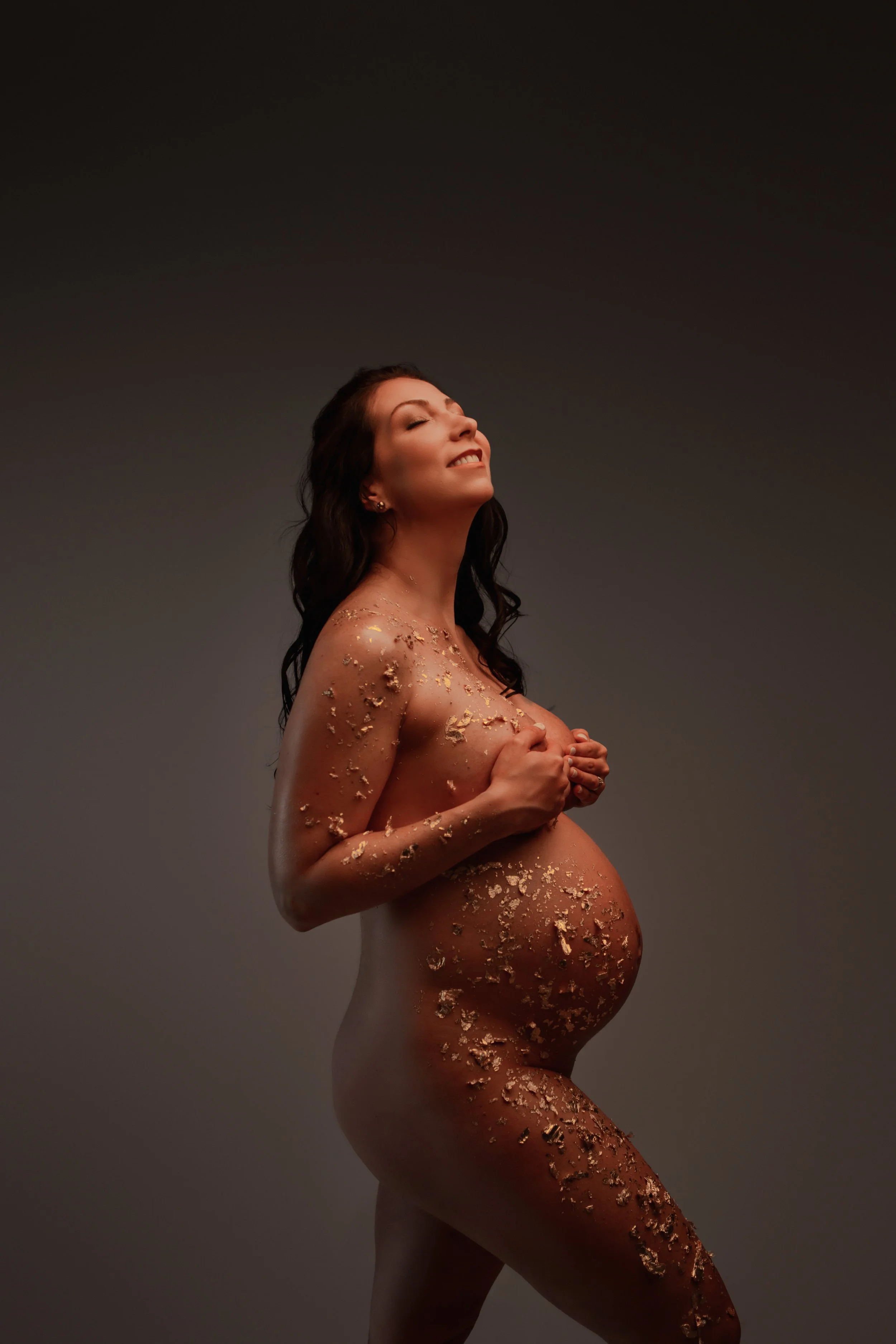 A nude pregnant woman covered in gold leaf, smiling with eyes closed, standing with hands on her chest against a gray background.
