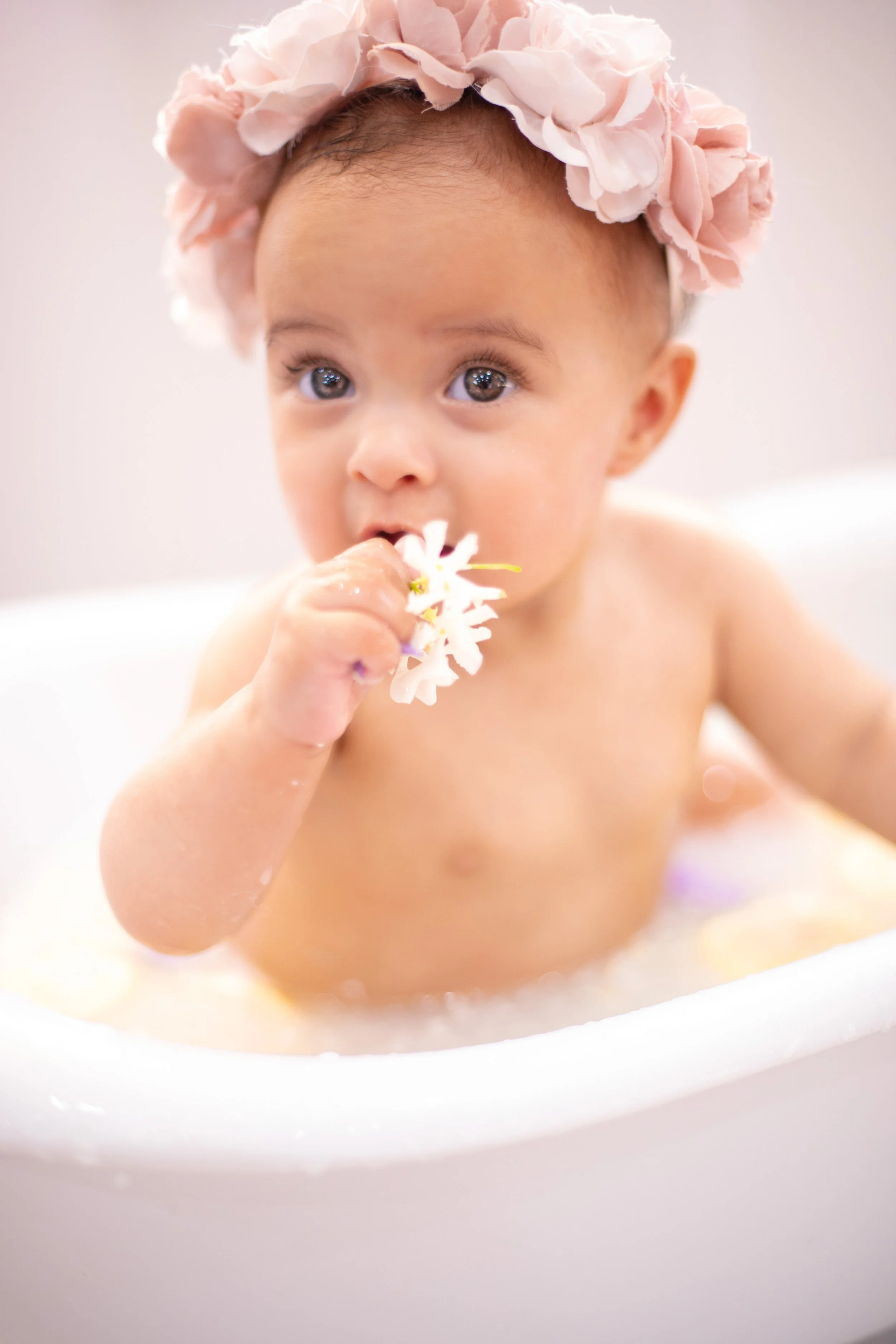 A young child with blue eyes and a pink flower crown drinks from a bath, holding a flower in their hand.