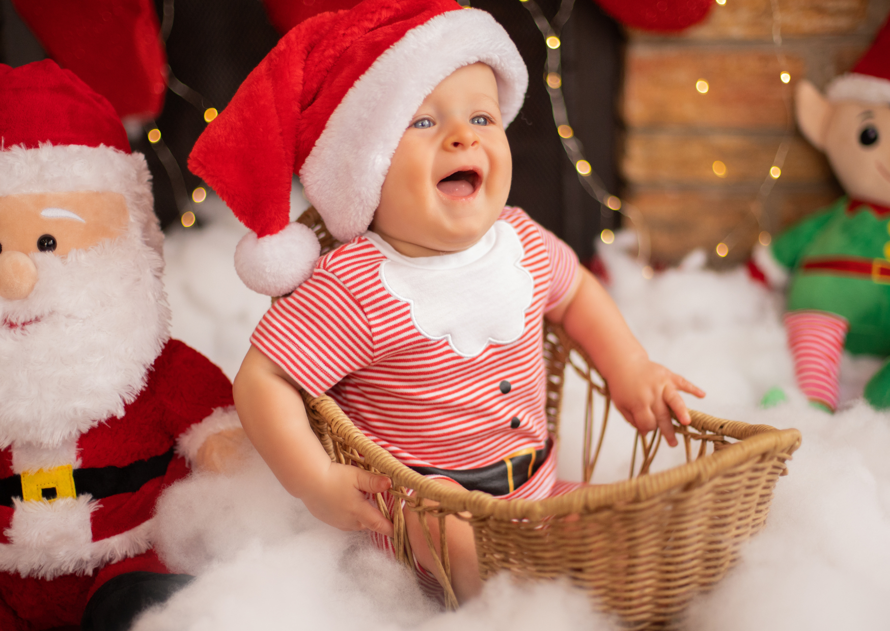 A baby wearing a red and white striped Christmas outfit and a Santa hat, sitting in a wicker basket, smiling amid Christmas decorations including Santa Claus and elf plush toys, with festive lights and snow-like cotton surrounding them.