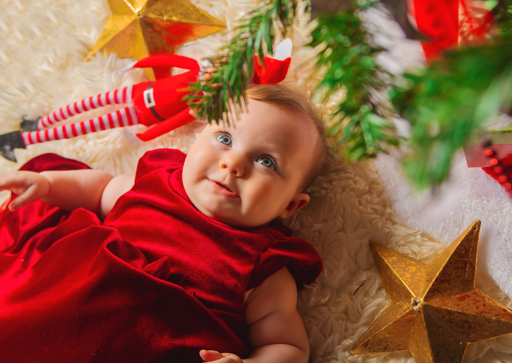 Baby in a red velvet dress lying on a fluffy white blanket surrounded by Christmas decorations, including a gold star, a Christmas tree branch, and an elf figurine with striped stockings.