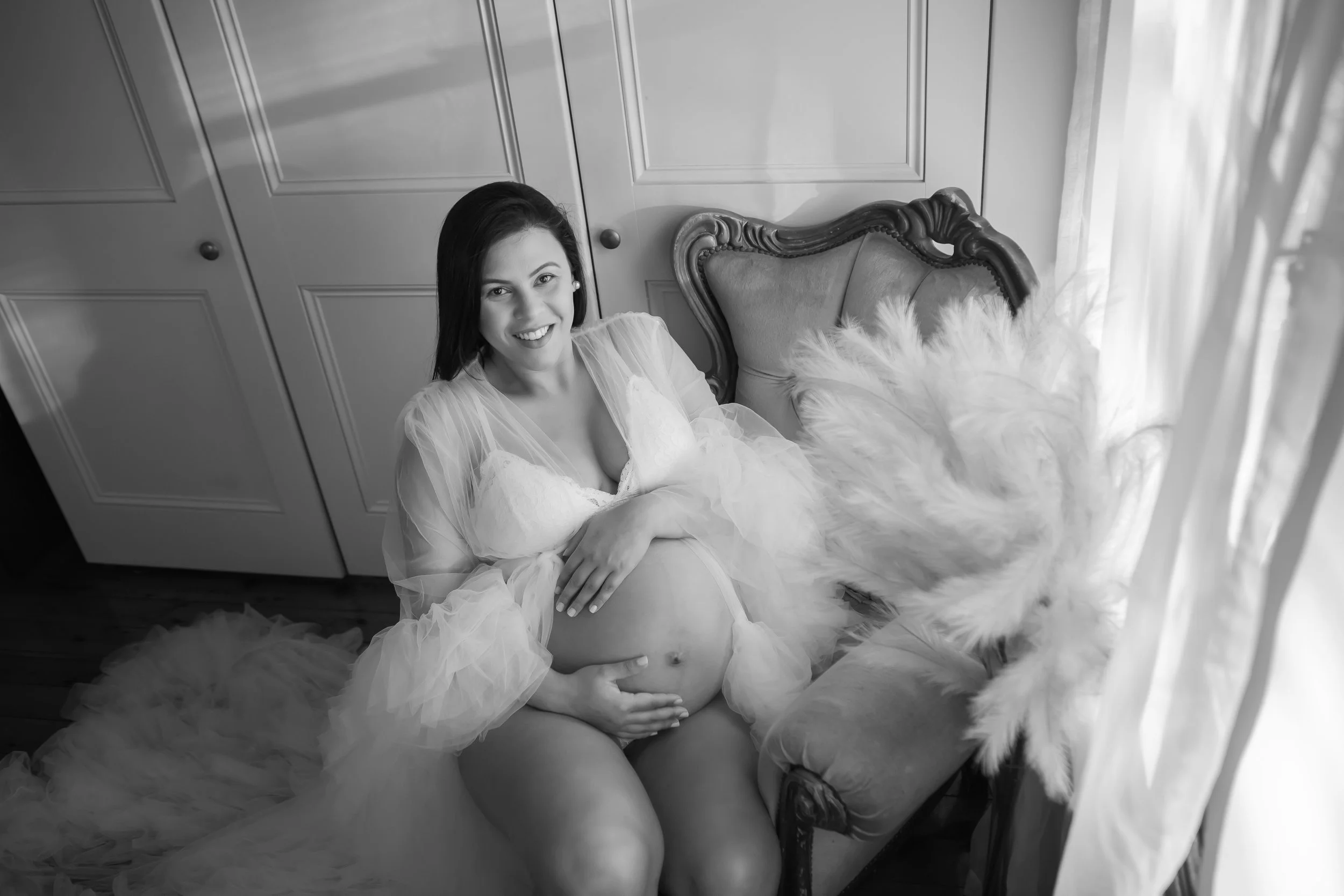 A pregnant woman with dark hair smiling, sitting on an elegant vintage chair with her dress open to show her belly, in a room with cabinet doors and sheer curtains, black and white photograph.