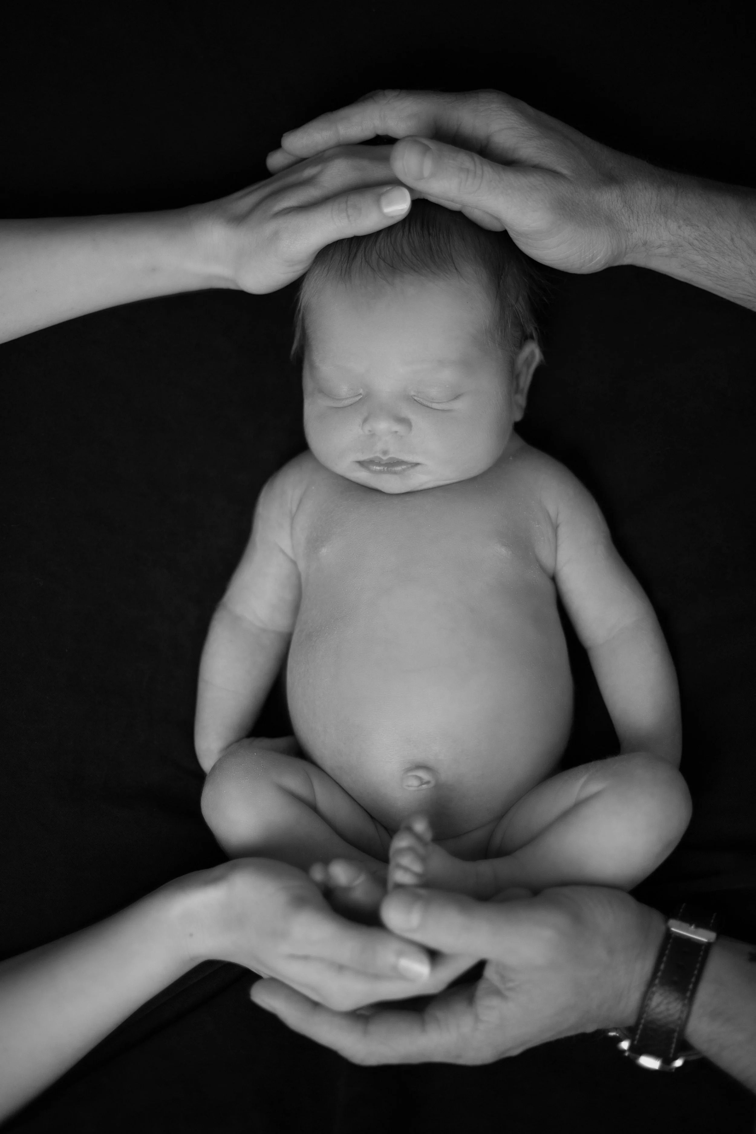 Black and white photo of a newborn baby with closed eyes, lying on a dark surface. The baby is surrounded by adult hands gently touching the head, feet, and main body.