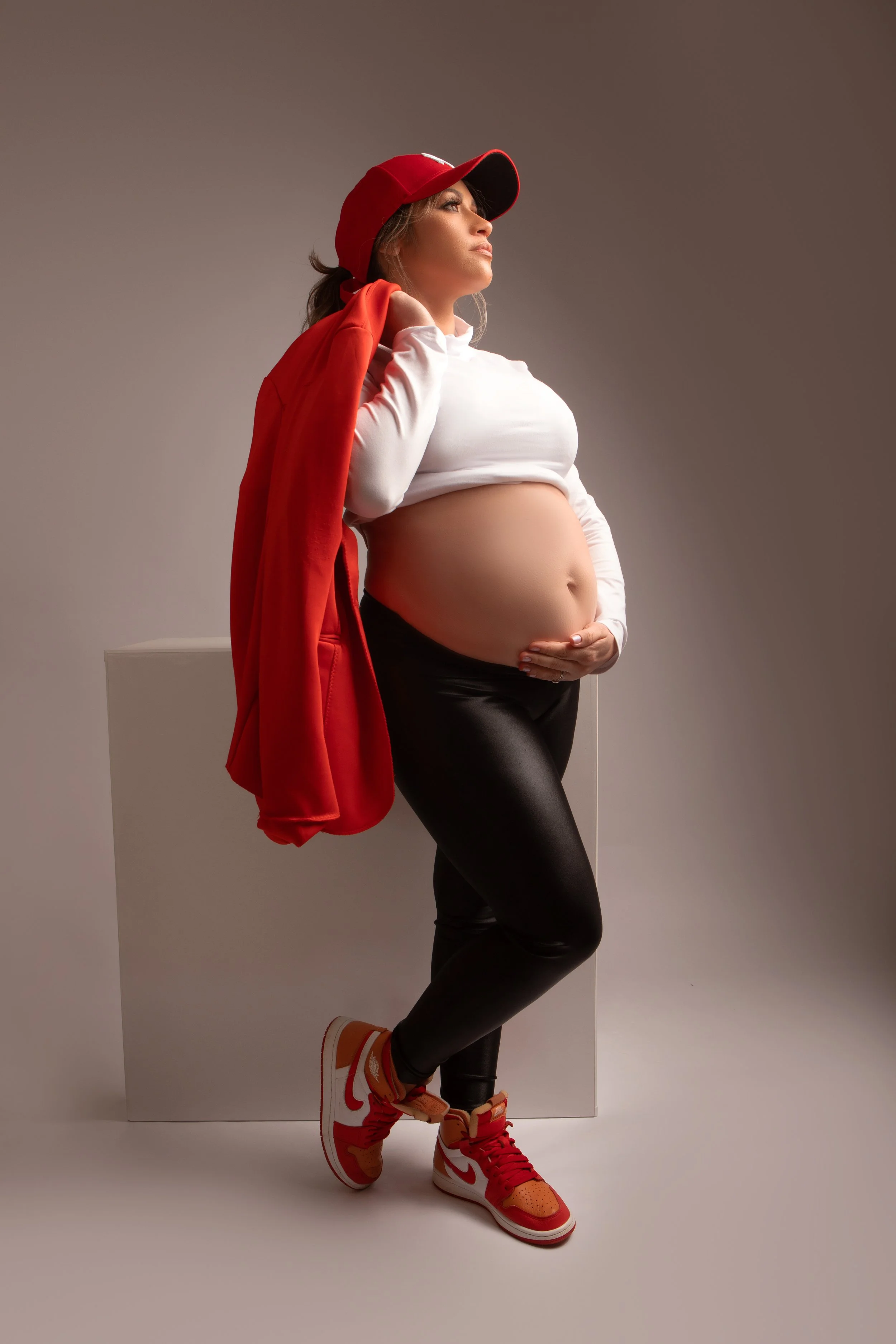 Pregnant woman wearing a red baseball cap, red jacket, white crop top, and black leggings, standing against a neutral background, holding her belly with one hand.