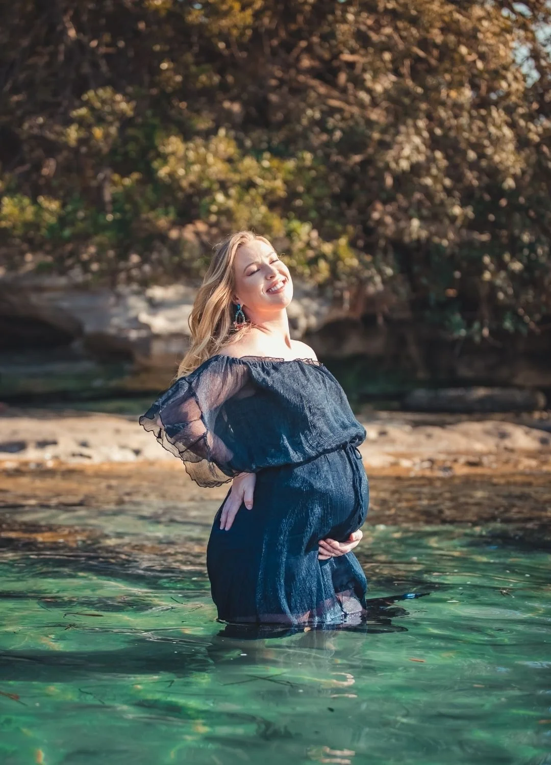 A pregnant woman standing waist-deep in clear water outdoors, smiling with her eyes closed, wearing a black dress with sheer sleeves, with a background of trees and rocks.
