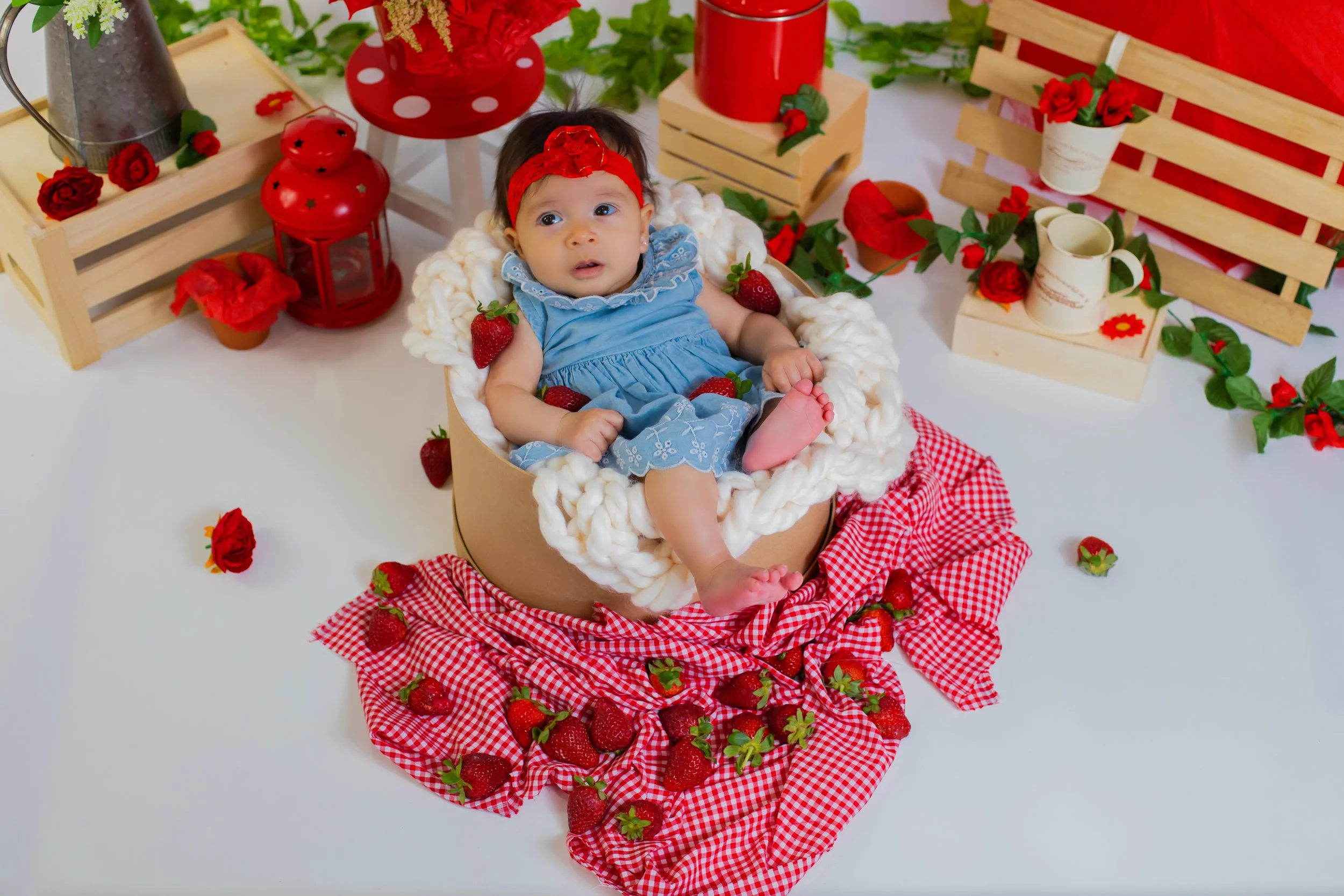 A baby girl in a blue dress sits in a round box surrounded by strawberries and red flowers. The background is decorated with wooden crates, red lanterns, potted flowers, and greenery, creating a festive, flower-themed setting.