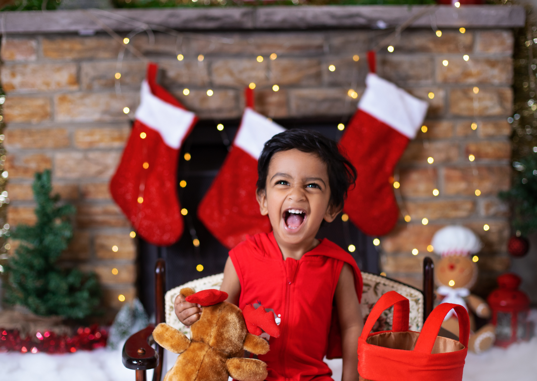 A young boy dressed in a red outfit sitting in a chair with Christmas stockings hanging on a brick fireplace behind him. He is holding a brown teddy bear and has his mouth open as if laughing or shouting. Christmas decorations, including small trees,