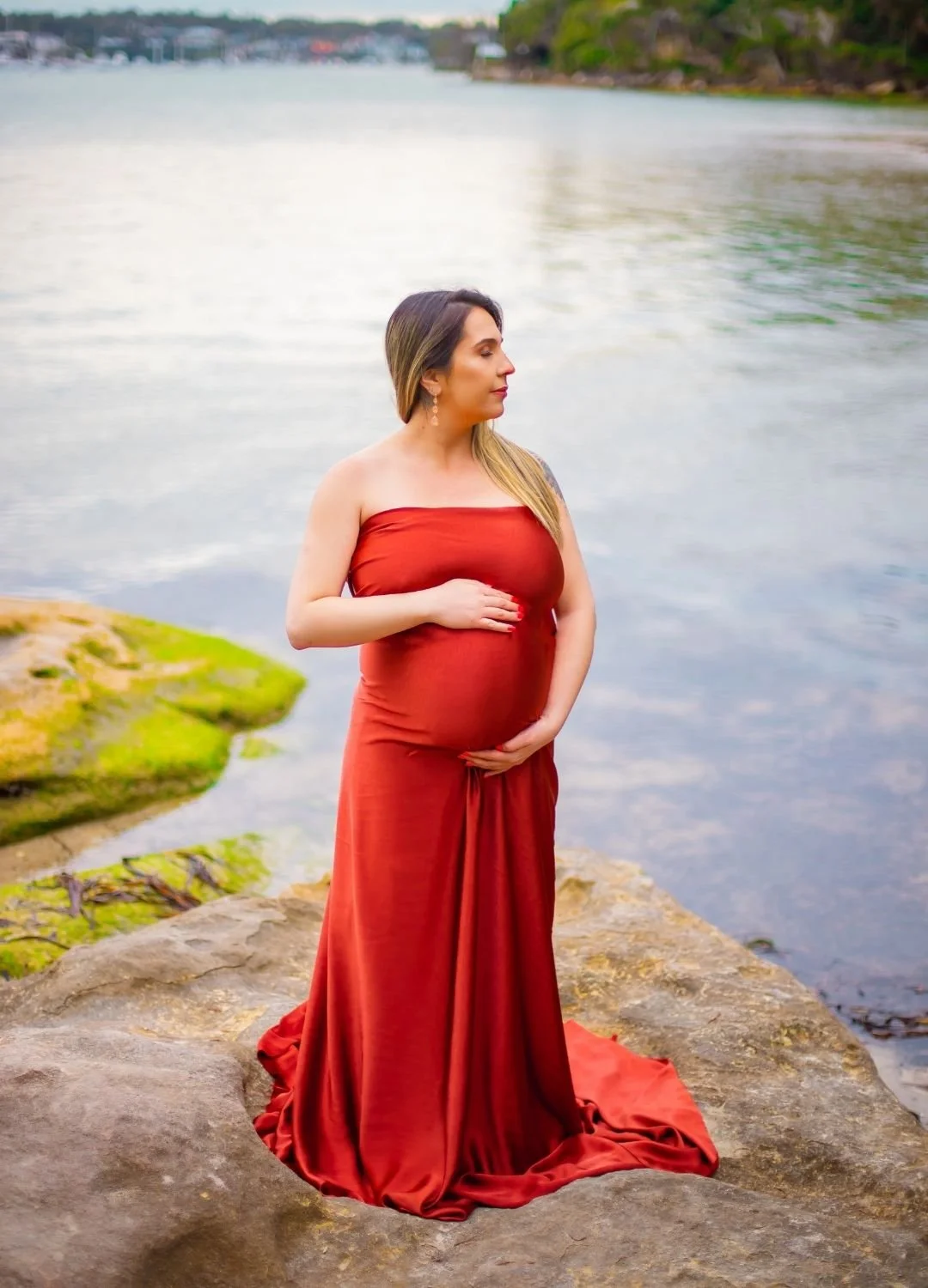 Pregnant woman in a red dress standing on a rocky shoreline by a body of water, with her hands resting on her belly, looking to the side.