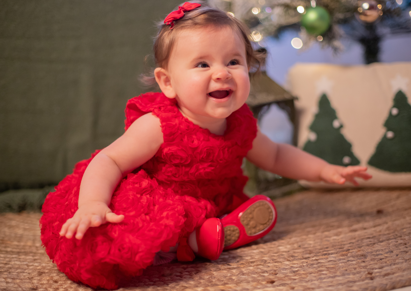 A cheerful young girl in a red dress with a red bow in her hair, sitting on a carpeted floor near a decorated Christmas tree and holiday-themed pillows.