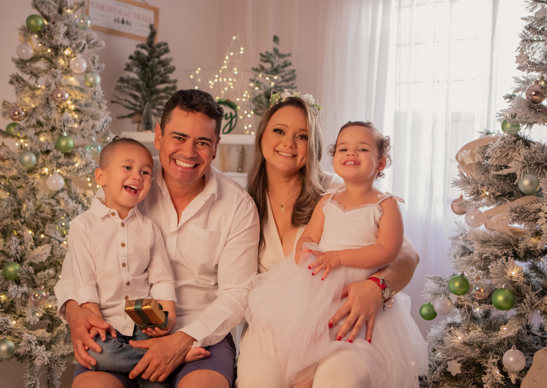 A family of four sitting together in front of decorated Christmas trees indoors. The father and son are on the left, the mother and daughter on the right, all smiling.