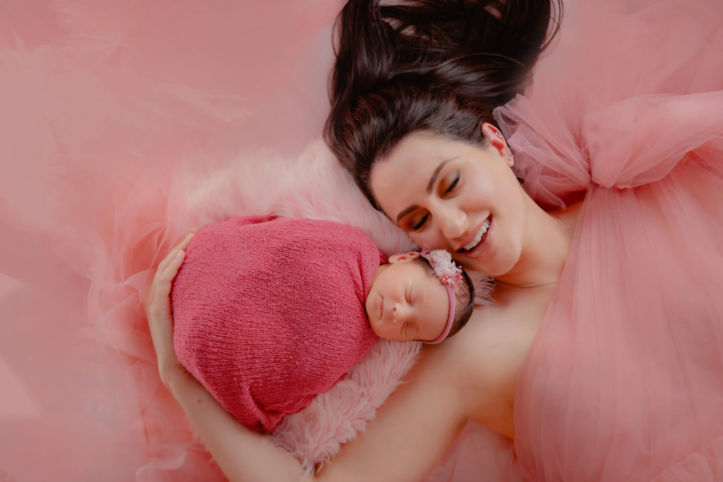 A woman lying on a pink fluffy blanket, smiling at a sleeping newborn girl wrapped in a pink cloth and wearing a pink headband with flowers.