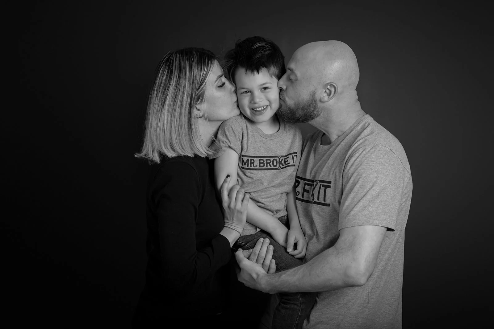A family of three with parents kissing their young son on both cheeks against a dark background.