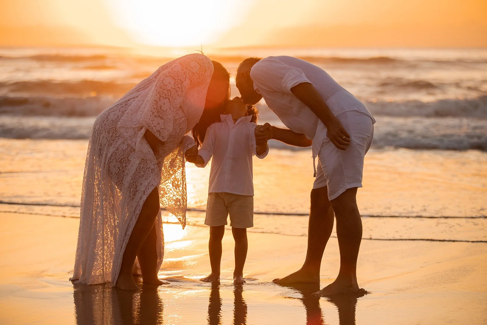 A family of three on the beach at sunset, holding hands and leaning in for a kiss.