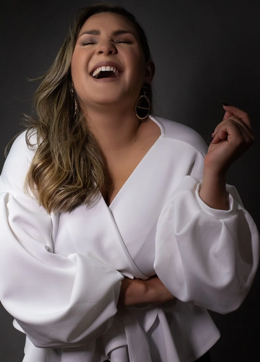 A woman with light brown wavy hair, wearing a white blouse, laughing with her eyes closed and hand raised, against a dark background.