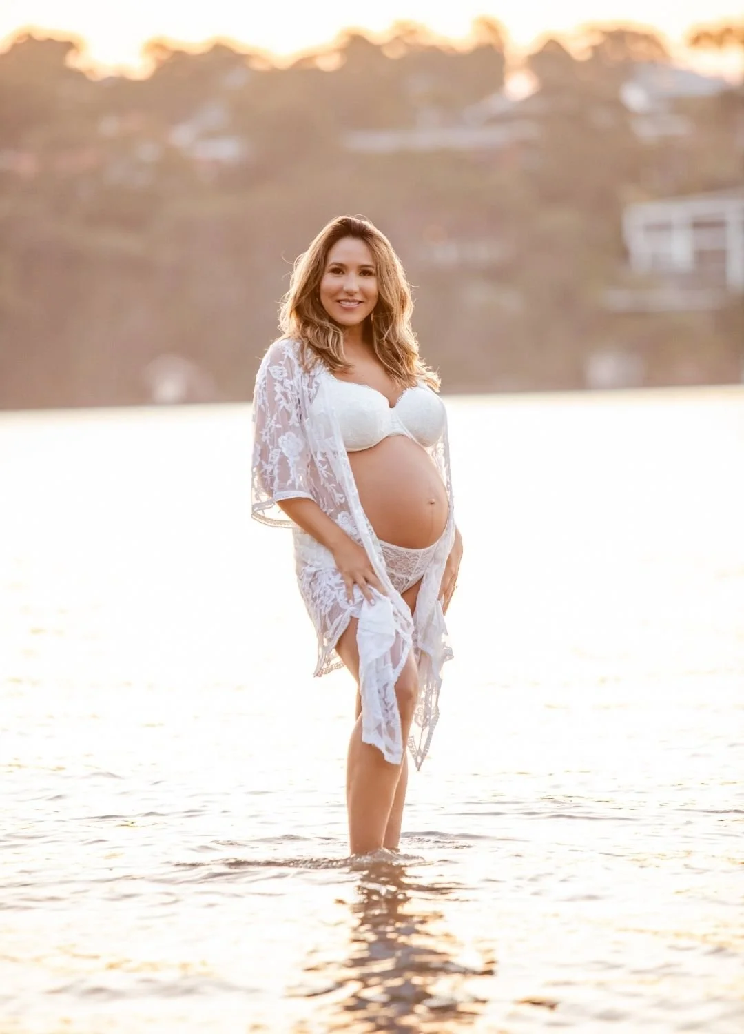 Pregnant woman standing in water during sunset, wearing white lace lingerie and a lace robe, smiling at the camera.