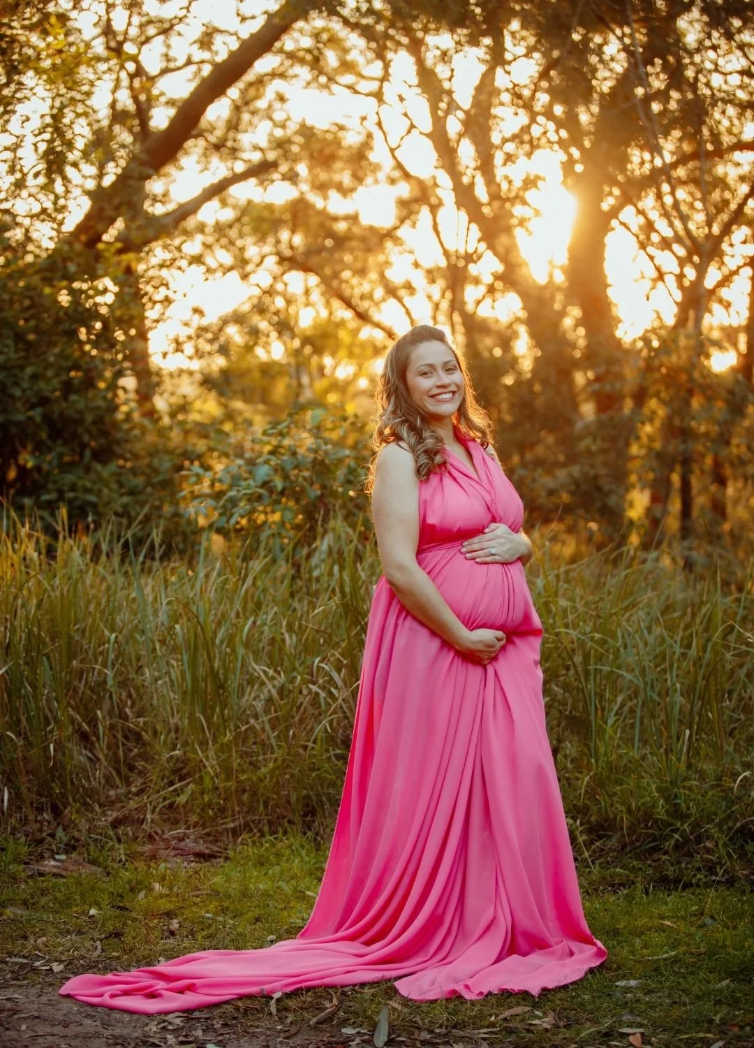 A pregnant woman in a flowing pink dress standing outdoors during sunset, smiling with trees in the background.