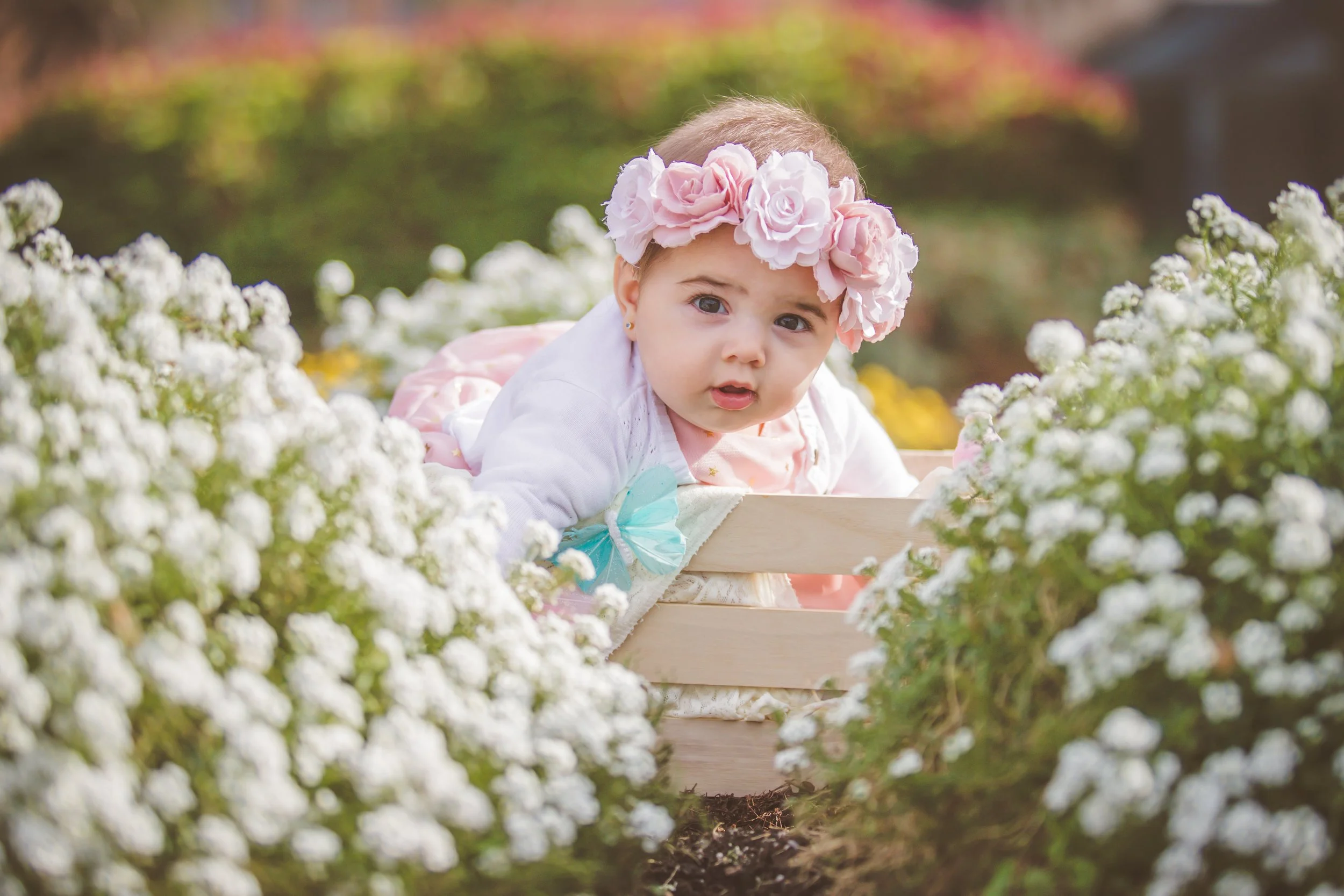 A young girl with a pink flower crown and a white cardigan crawling on a wooden box surrounded by white flowers in a garden.