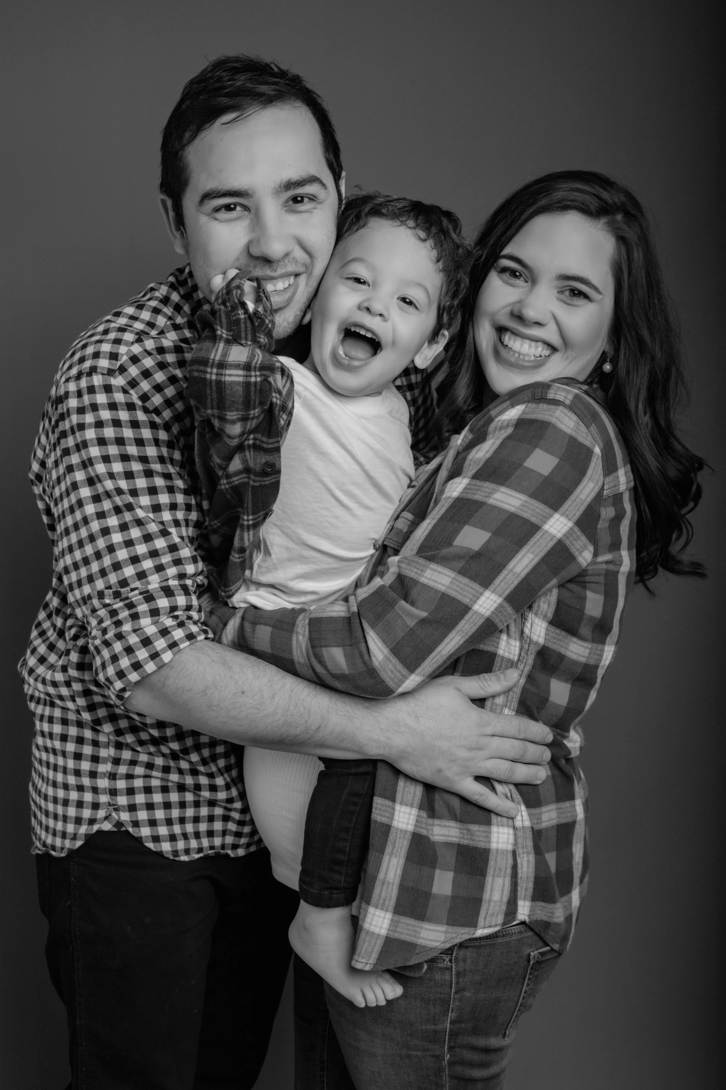 A happy family of three smiling and hugging each other in a studio. The father, mother, and young child are in a close embrace, all wearing plaid shirts. The photo is in black and white.