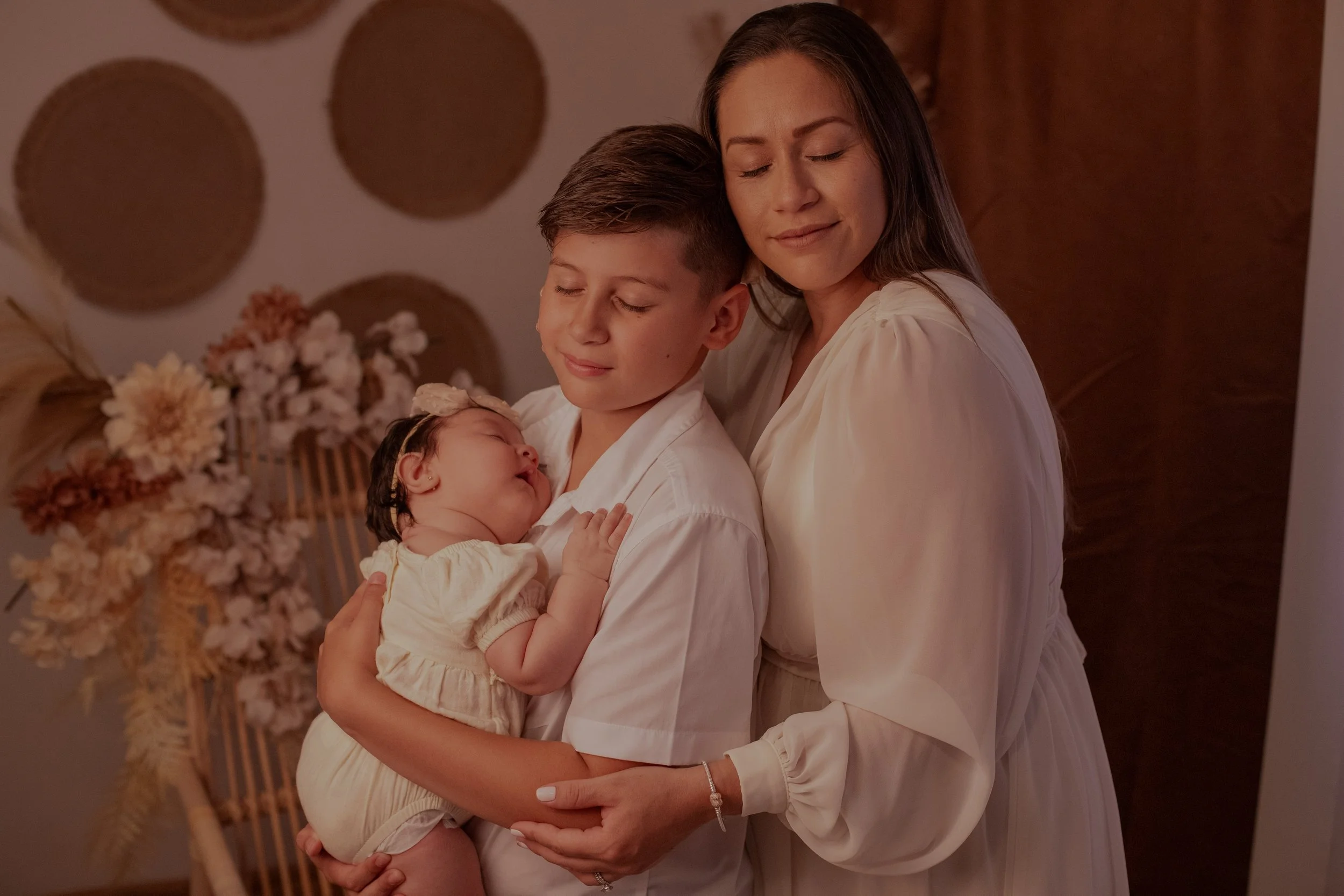 A woman holding a sleeping baby in her arms, with a boy standing beside her, also holding the baby, all with peaceful expressions.