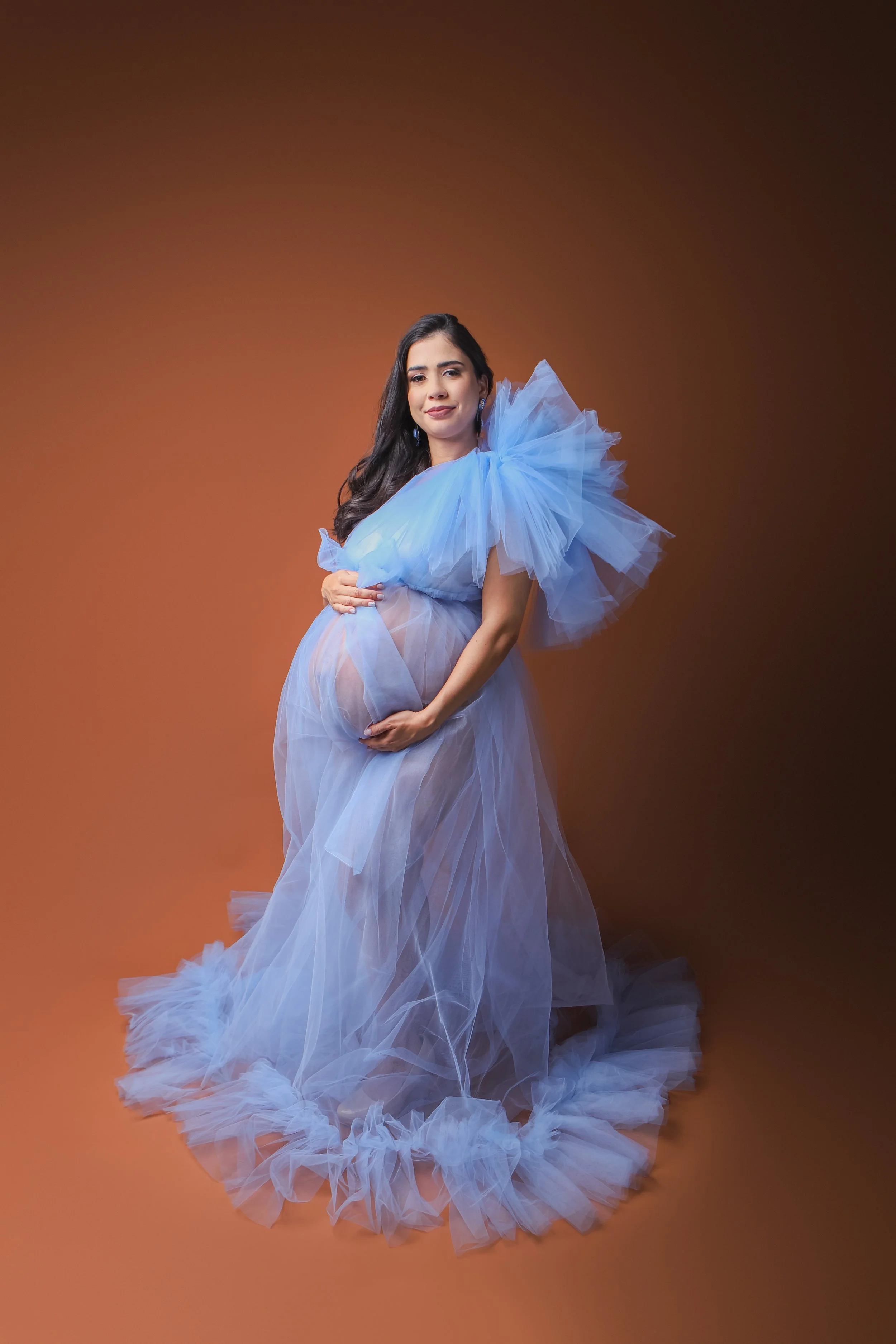 A pregnant woman in a flowing, sheer blue tulle gown with ruffled sleeves standing against a brown gradient background, gently holding her belly with a slight smile.