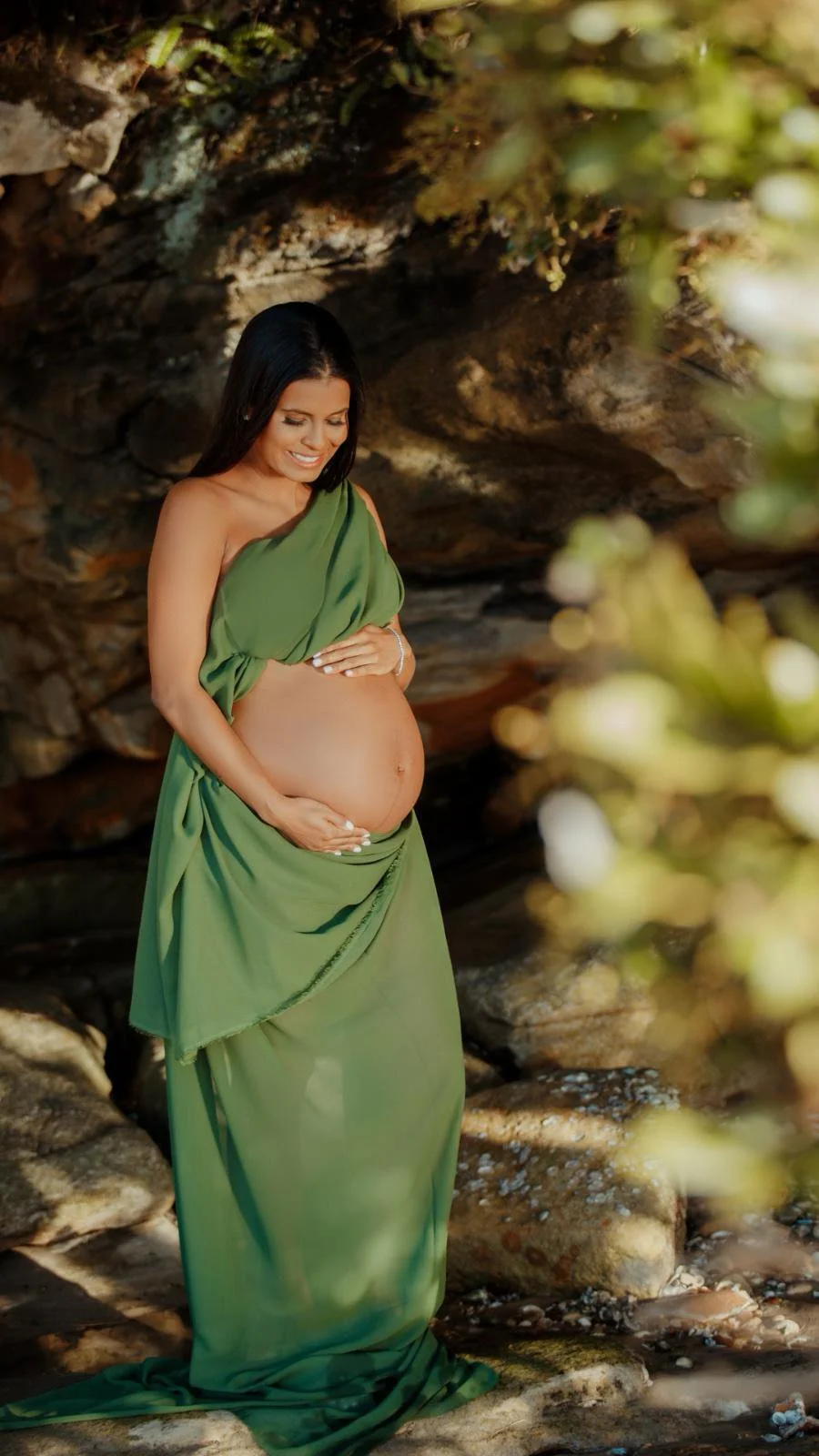 Pregnant woman in a green dress standing in a rocky outdoor setting, smiling and touching her belly.