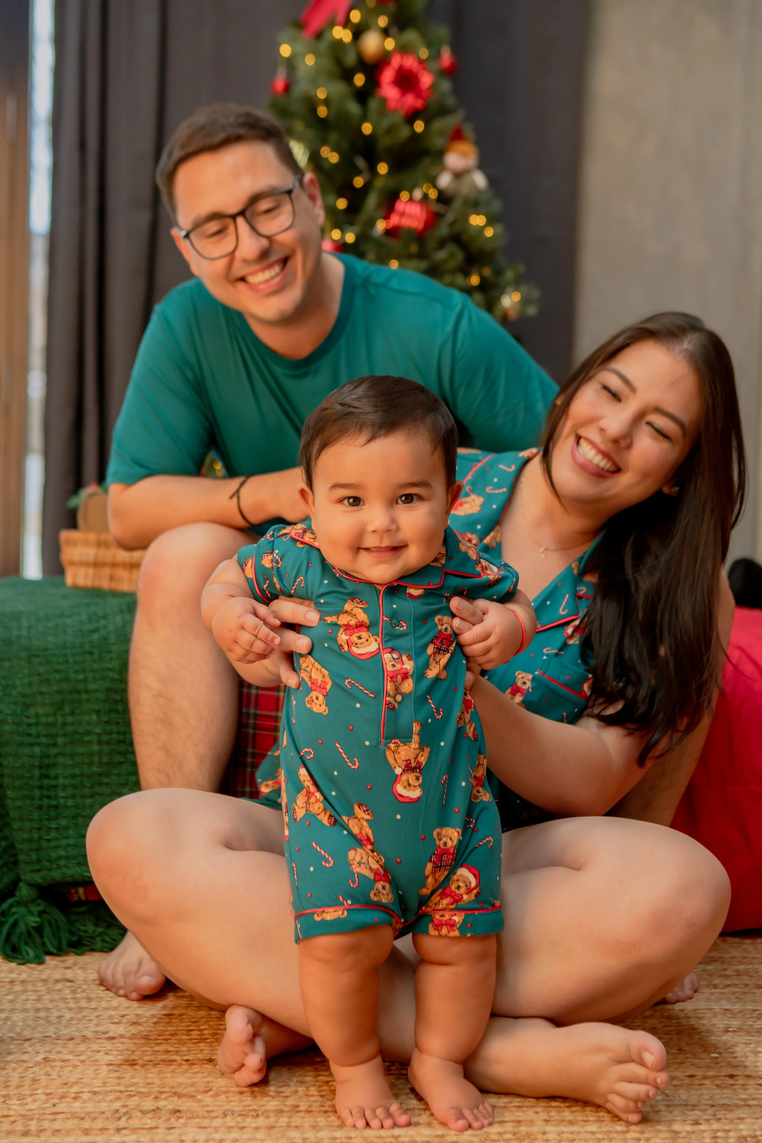 A happy family of three celebrating Christmas indoors; a woman and man are sitting on the floor holding their smiling toddler, with a decorated Christmas tree in the background.