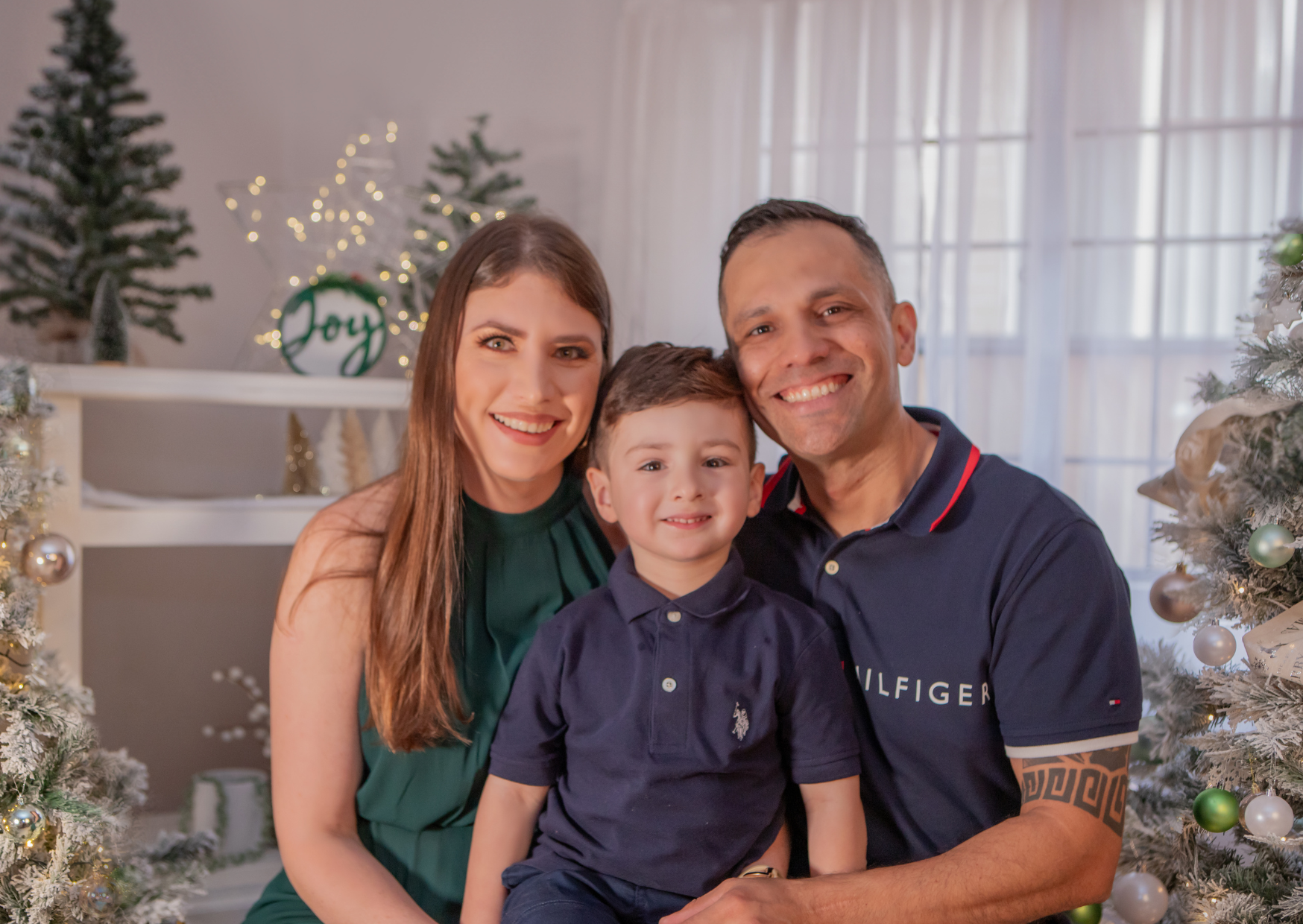 A smiling family of three, including a woman, a man, and a young boy, poses together in front of decorated Christmas trees with ornaments and lights, near windows with white curtains.