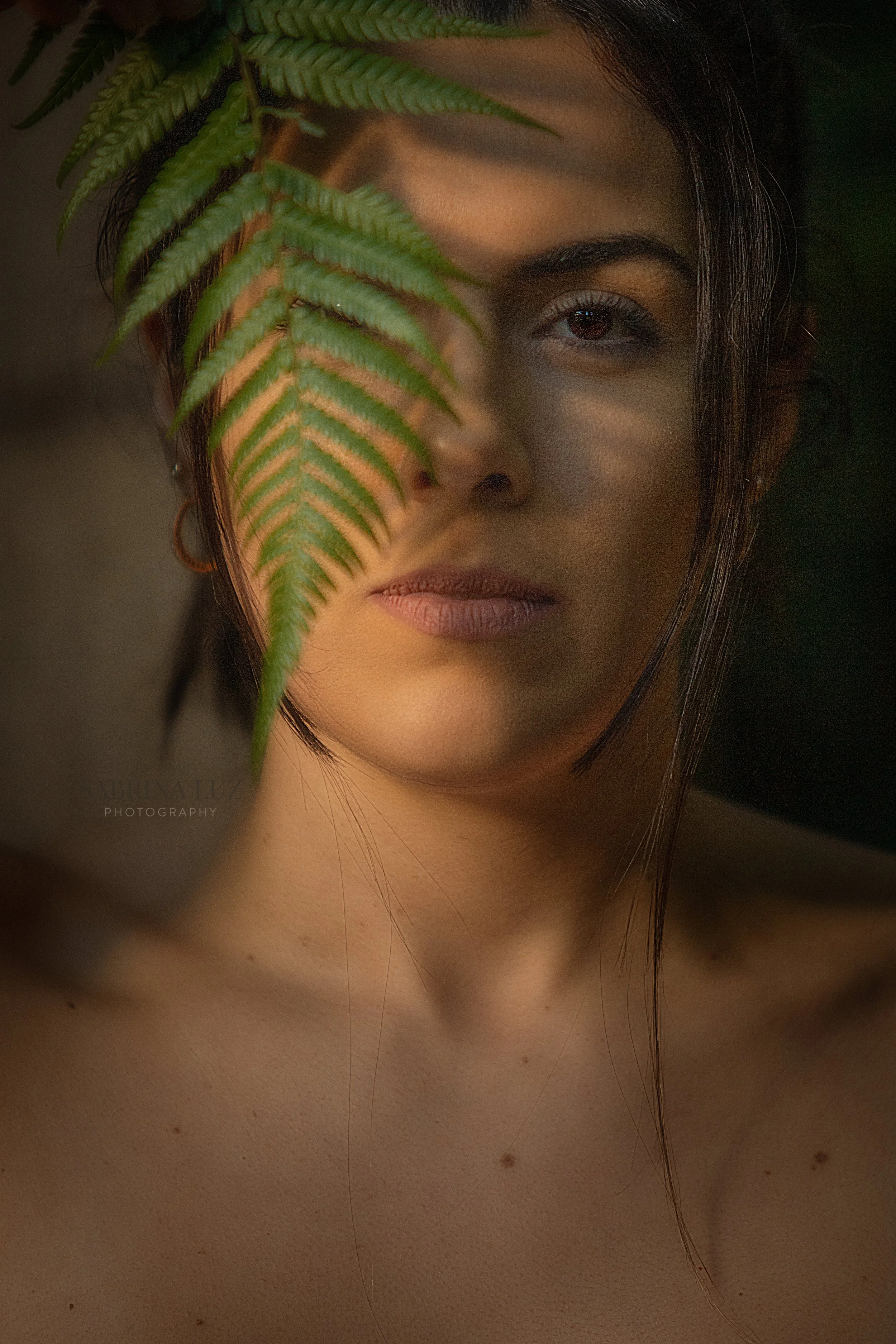 A woman with dark hair and brown eyes partially obscured by a large green fern leaf, with warm lighting on her face and upper shoulders.