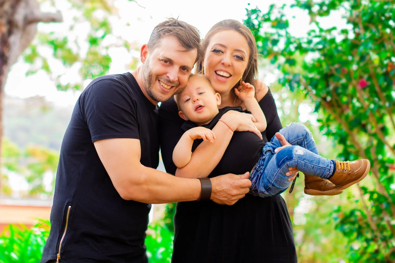 Happy family of three, a man, a woman, and a toddler, outdoors with green trees in the background. The man and woman are smiling and holding the toddler.