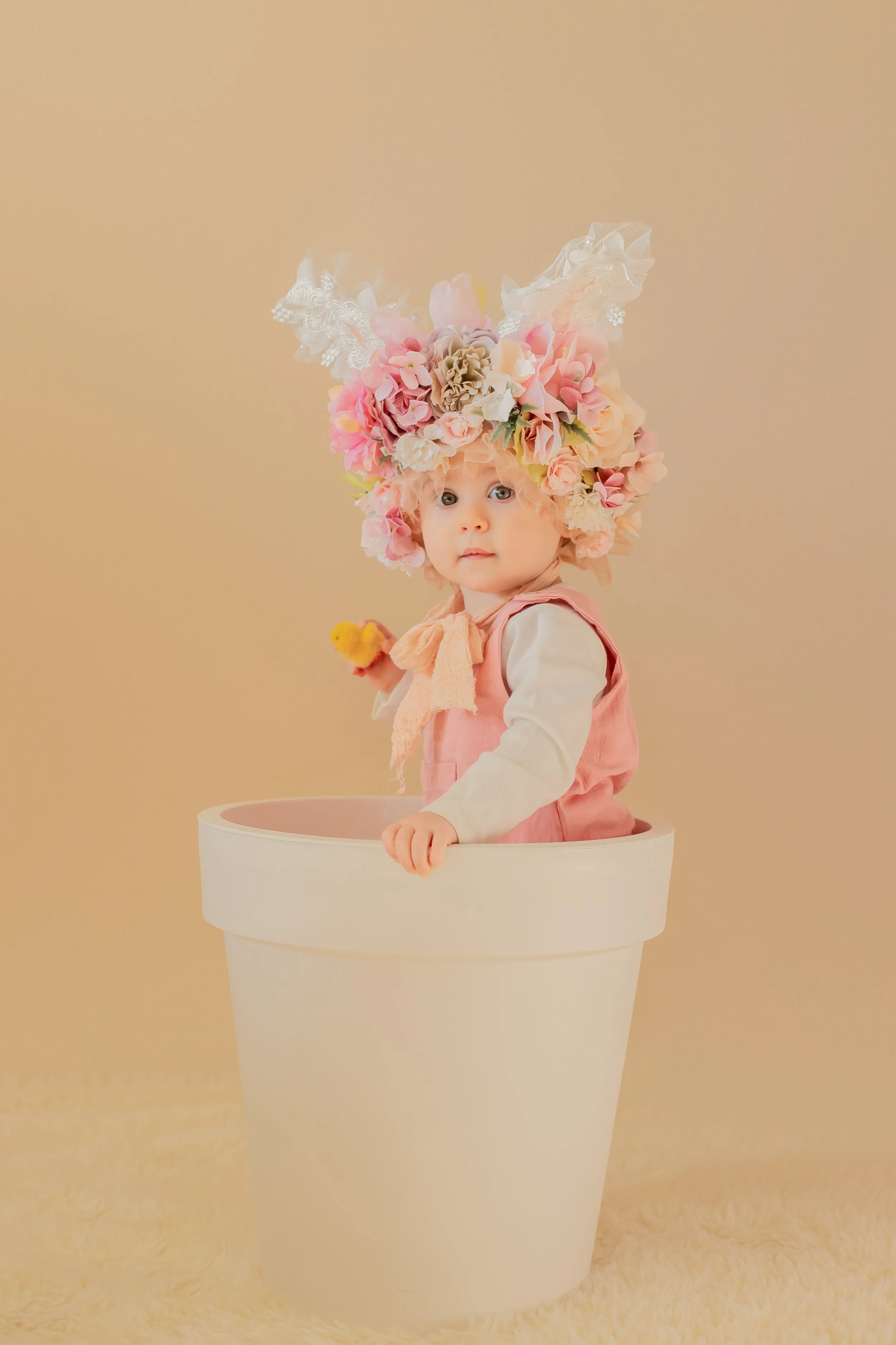 Baby wearing a large flower headdress inside a white flowerpot, holding a small yellow toy duck, against a beige background.
