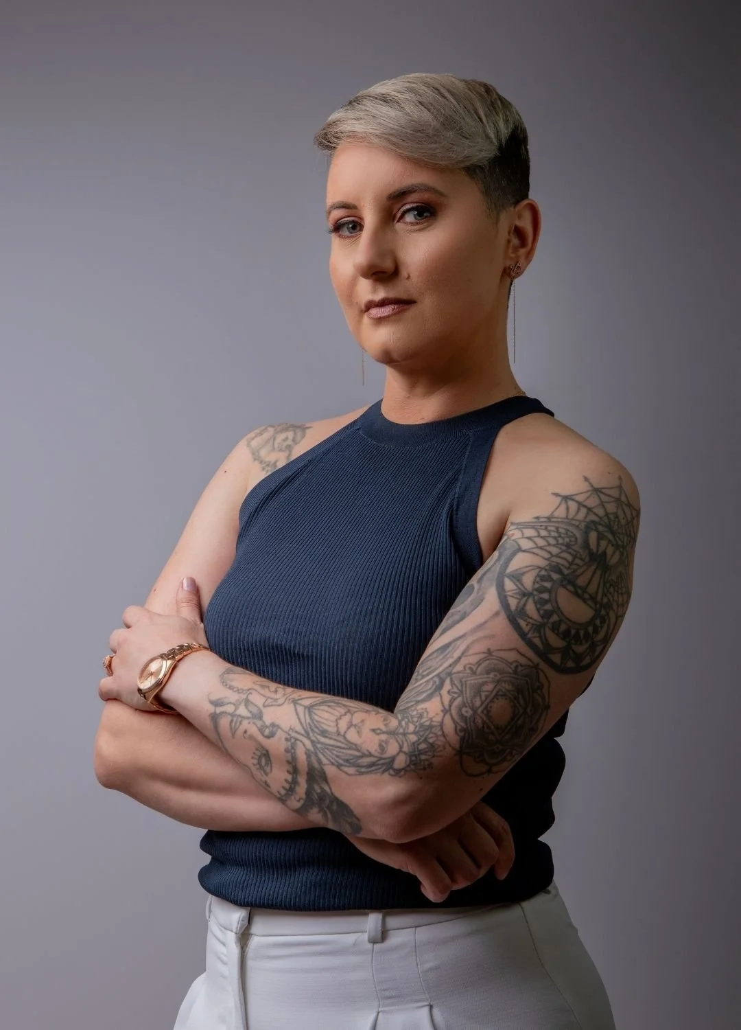 A woman with short, blonde hair, wearing a navy sleeveless top and white pants, crossed arms, and tattoos on both arms, standing against a plain gray background.