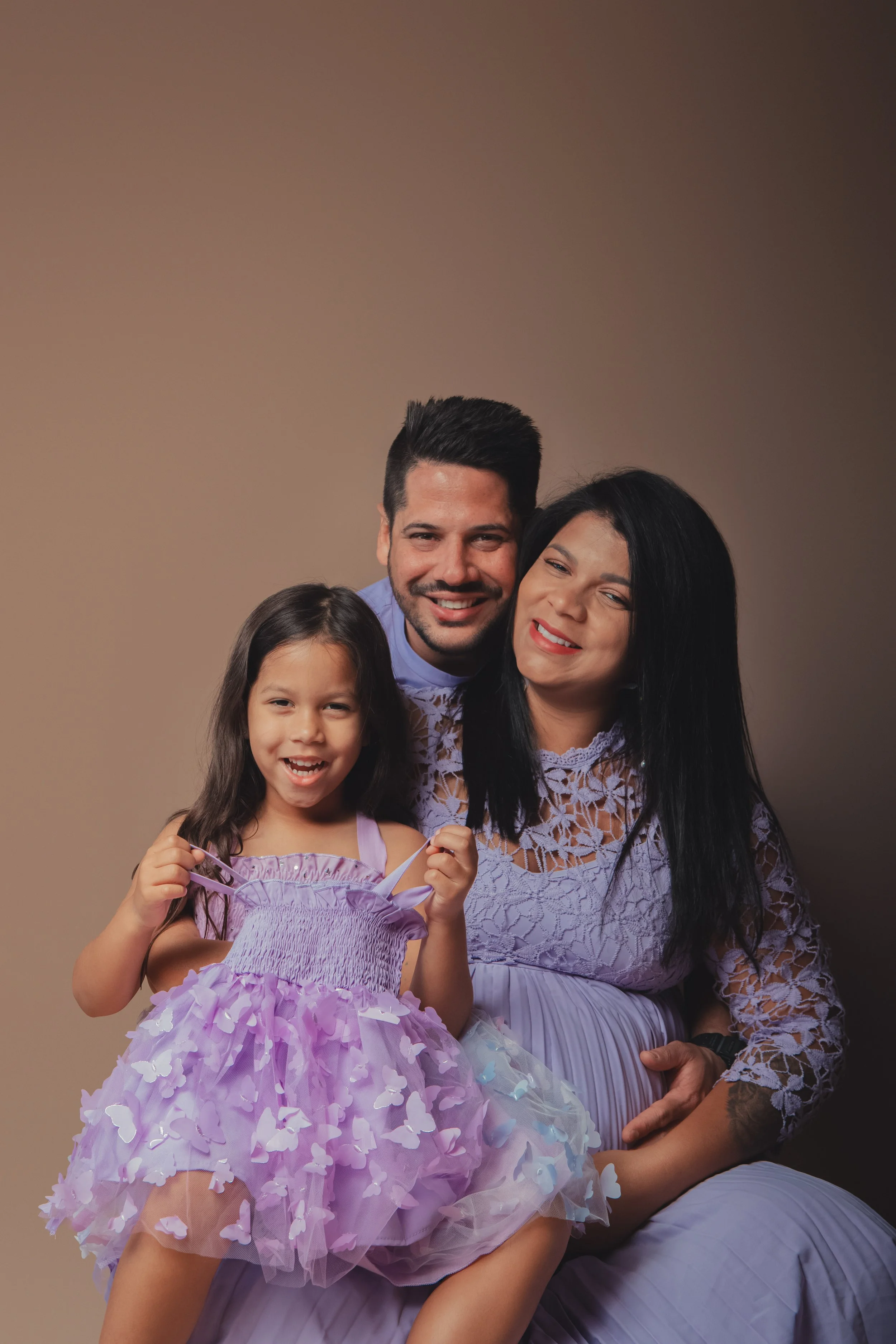 A smiling family of three, including a man, woman, and young girl, dressed in lavender clothing against a neutral background.