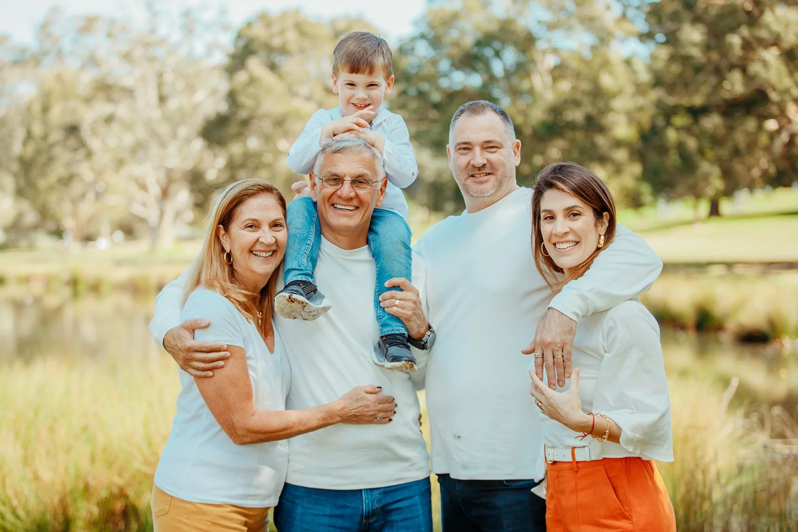 A smiling family of six posing outdoors in a park, with trees and a pond in the background. They are dressed casually, with one young boy sitting on an older man's shoulders.