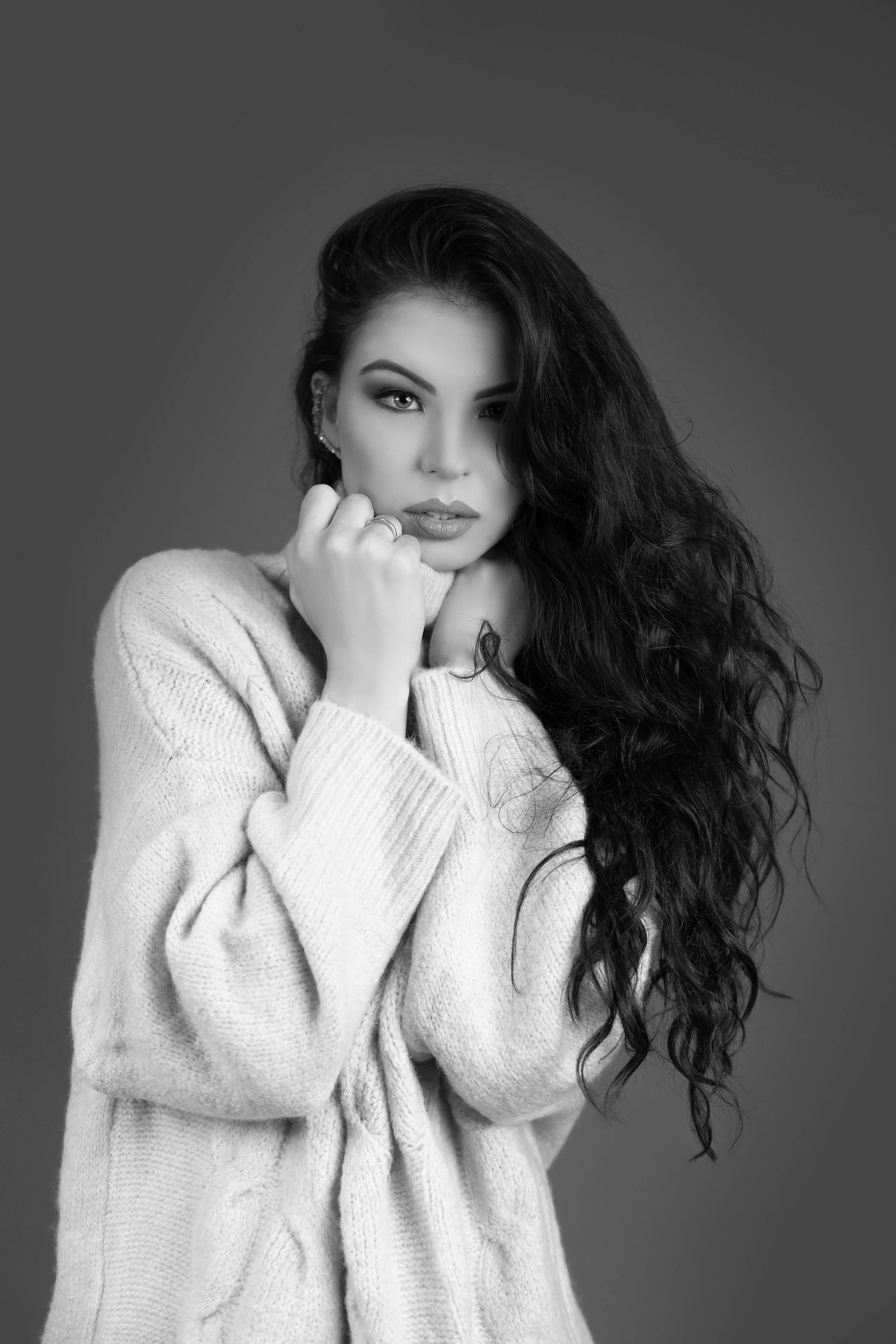 Black and white portrait of a woman with long curly hair, wearing a chunky knit sweater, looking directly at the camera with a thoughtful expression, one hand resting on her chin.